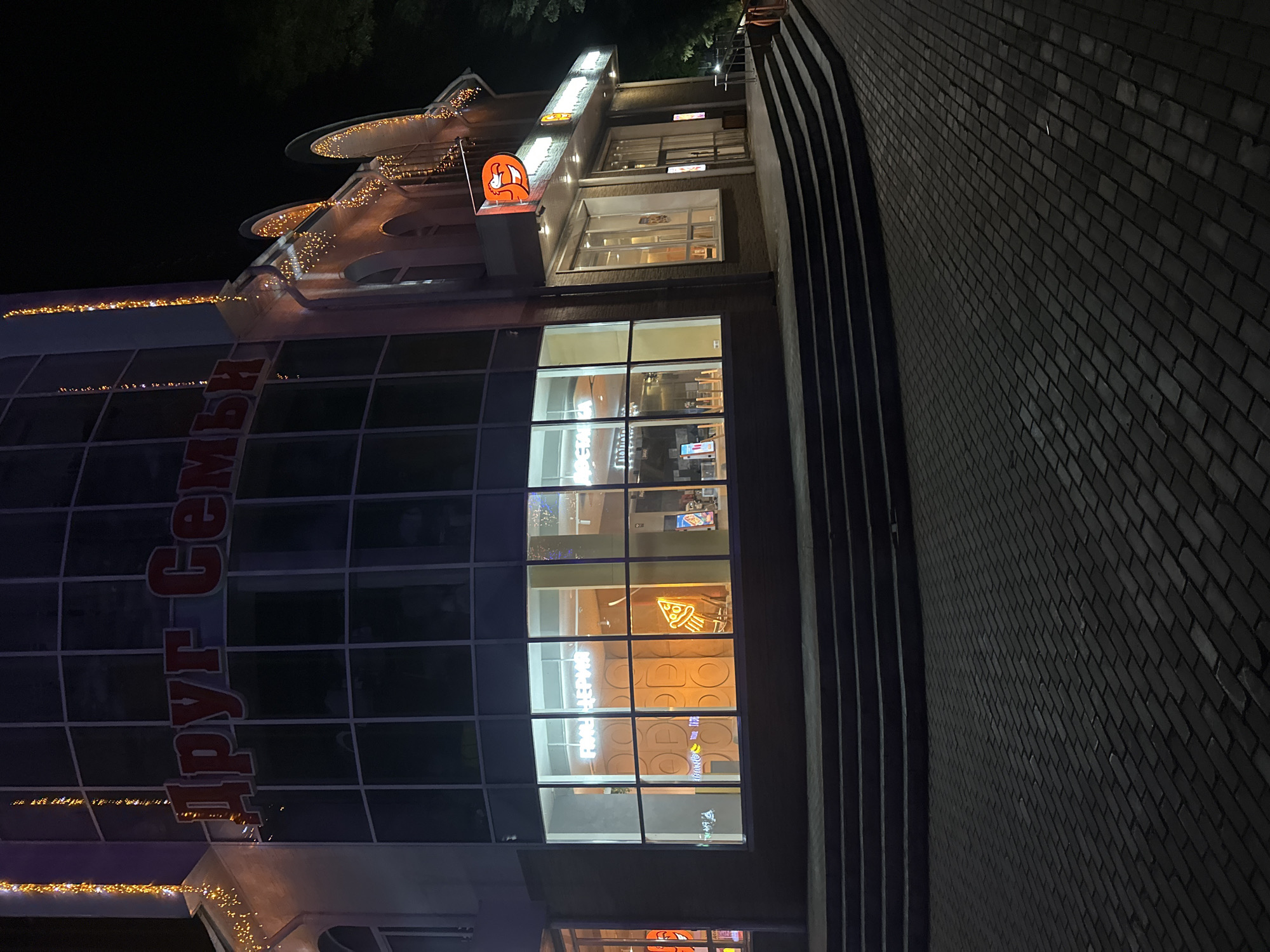 This is a nighttime, eye-level, wide shot of a brick building with large windows, adorned with string lights. The building has a modern design with curved architectural elements on the upper floors. The scene is illuminated by the building's interior lights, neon signs, and decorative string lights. The overall mood is festive and inviting.
On the left side of the building, large red Cyrillic lettering is visible, spelling "Друг Семьи" which translates to "Friend of the Family." Above this, a neon sign with an orange fox-like character is illuminated.
Through the large windows, the interior of the building is visible, revealing brightly lit spaces with some displays and furniture. In one window, a neon sign depicting a slice of pizza is prominently displayed. The upper floors also have illuminated signs, though the text is less clear.
To the right of the building, a dark brick wall forms a pathway or staircase, also illuminated by the building's lights. The sky is dark, indicating nighttime. The weather appears calm, with no visible rain or wind.
The image captures a lively urban setting, likely a commercial establishment or entertainment venue, in Birobidzhan, Russia. The combination of interior lighting, neon signs, and festive string lights creates a warm and welcoming atmosphere.