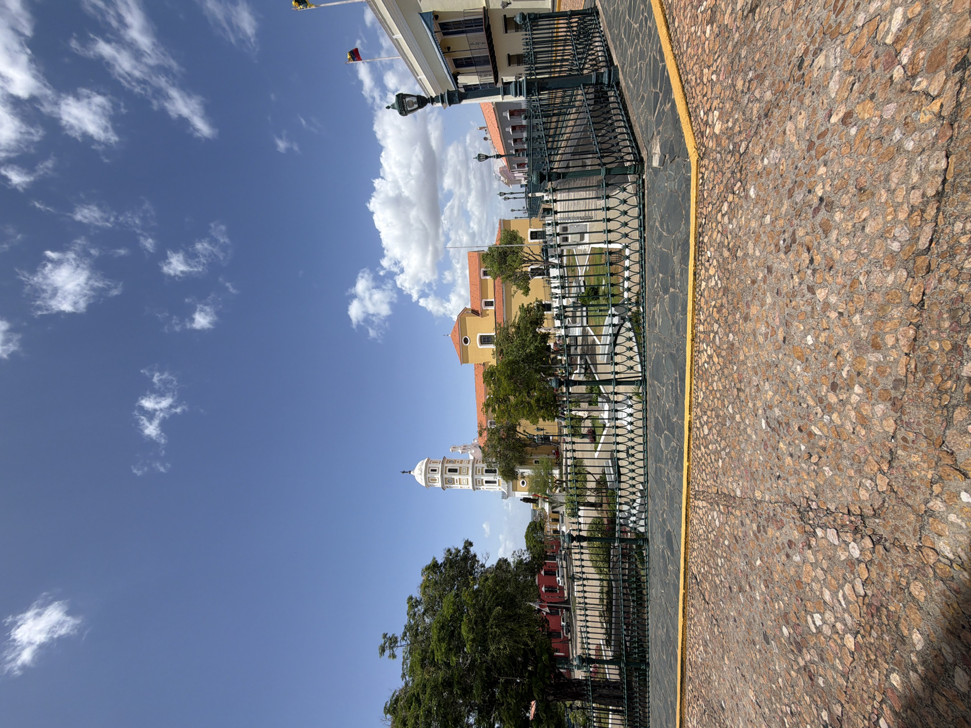 This photograph depicts a street scene in Ciudad Bolívar, Venezuela, with a clear, blue sky dotted with scattered clouds above. The view is framed by a decorative wrought-iron fence along the lower edge of the image and a cobblestone pavement on the right. Beyond the fence, there is a paved street with buildings of varying colors and architectural styles. A prominent white church tower stands out against the sky. Trees with green foliage are visible in the background. Flags are visible on top of some of the buildings. The image captures a tranquil, sunny day in this urban setting.