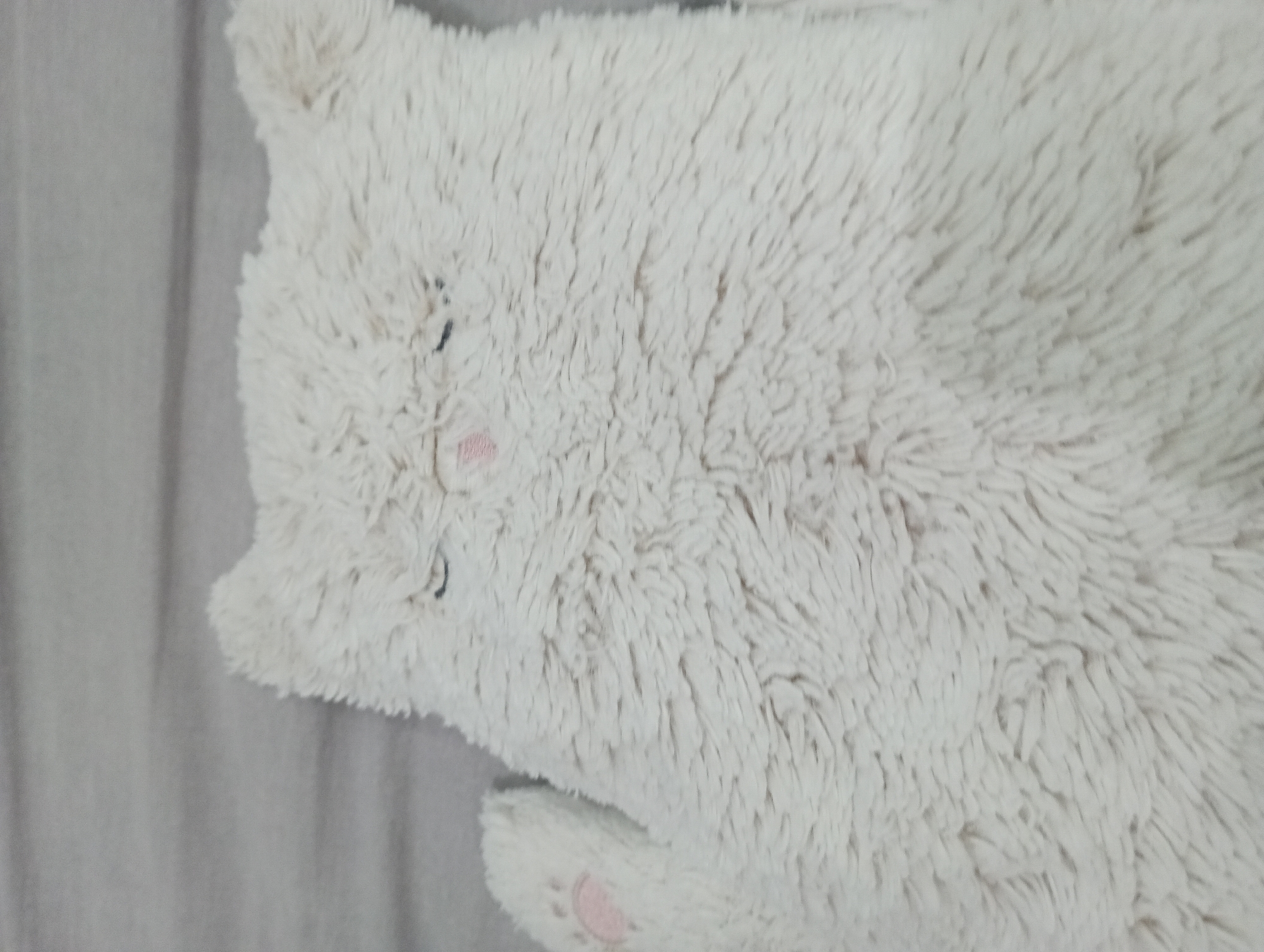 The image depicts a plush white stuffed animal, likely a bear or similar creature, laying on a gray surface. The toy has a soft, fuzzy texture and simple facial features: two black stitched eyes and a pink stitched nose. One paw is visible and shows a pink paw pad. No people are present. The context of Nha Trang Ward, Vietnam, is not discernable.