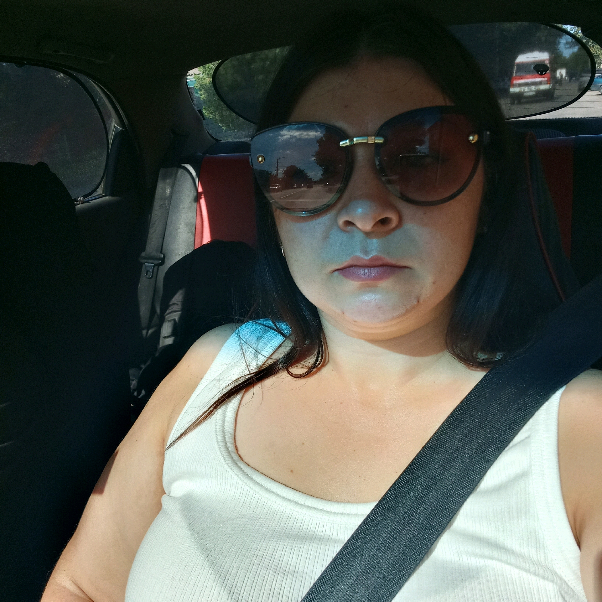 A woman with dark hair and wearing large sunglasses and a white tank top is pictured inside a car. She is wearing a seatbelt. The interior of the car is visible, including the back seat and window shades. Outside the car, a road and vehicles are visible through the window. The image appears to be taken from the perspective of the person in the passenger seat. The environment suggests a daytime setting, likely a road trip or commute. There are no visible indications of the specific location within Kropyvnytskyi, Ukraine.