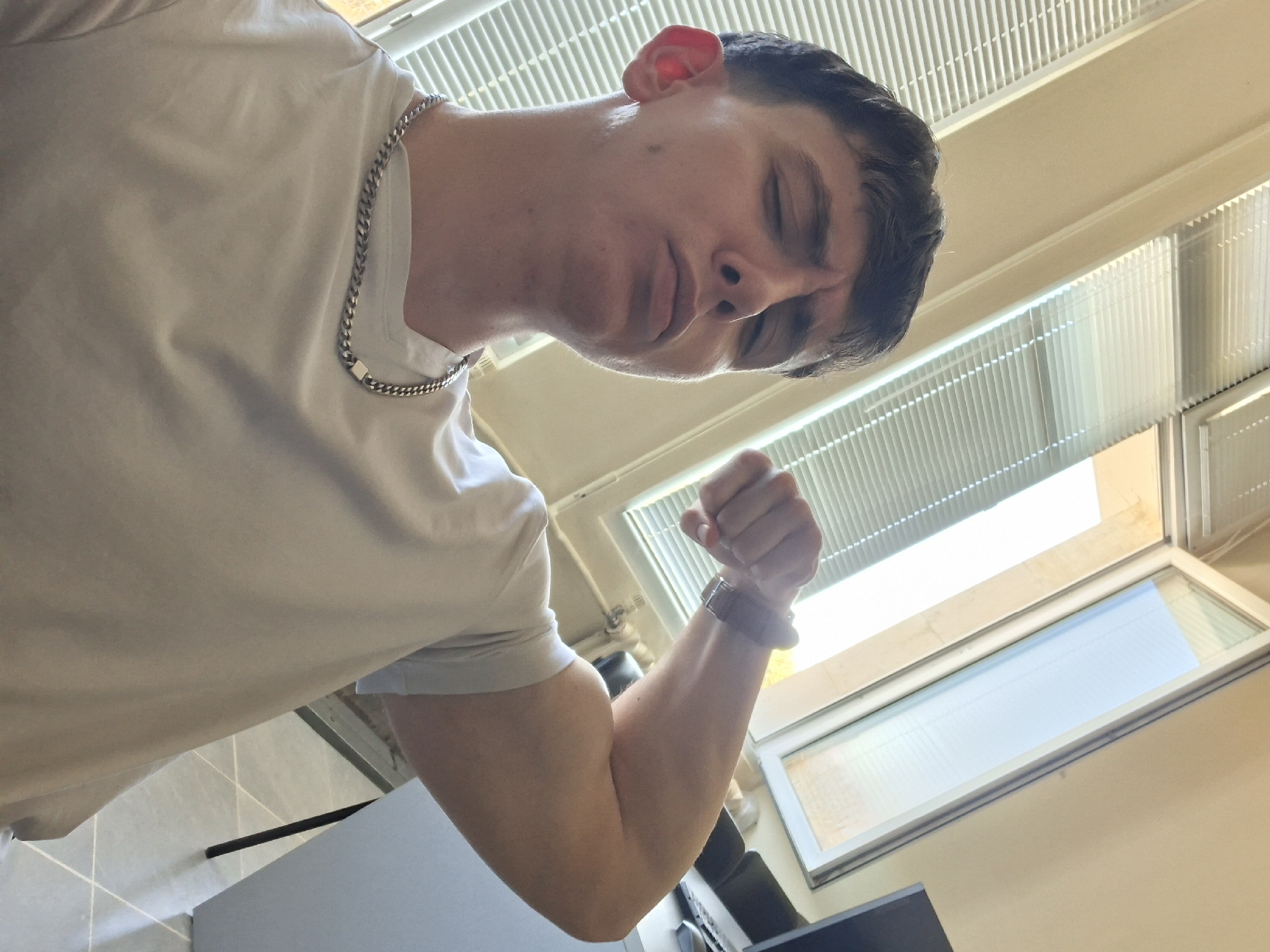 A man wearing a white t-shirt and a silver chain is flexing his arm. He is wearing a watch on his wrist. The background includes blinds and a window. The location is Unknown, Unknown.
