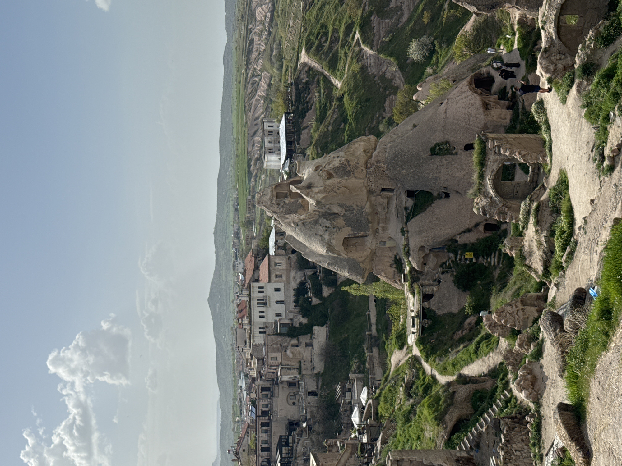 This is a vertical, high-angle shot in Uçhisar Beldesi, Turkey, showing a landscape dotted with historical stone structures and dwellings. The sky is pale blue with white clouds on the left.
In the foreground, a pathway leads up to the right, populated by a few people walking and looking around. Further down, buildings and dwellings built into the rock formations are noticeable. Some of the rock formations have openings and windows, showing their use as living spaces.
The middle ground presents a dense cluster of buildings and houses that are close together. The area is somewhat hilly and vegetated.
In the background, a larger vista of green-covered hills stretches towards the horizon.