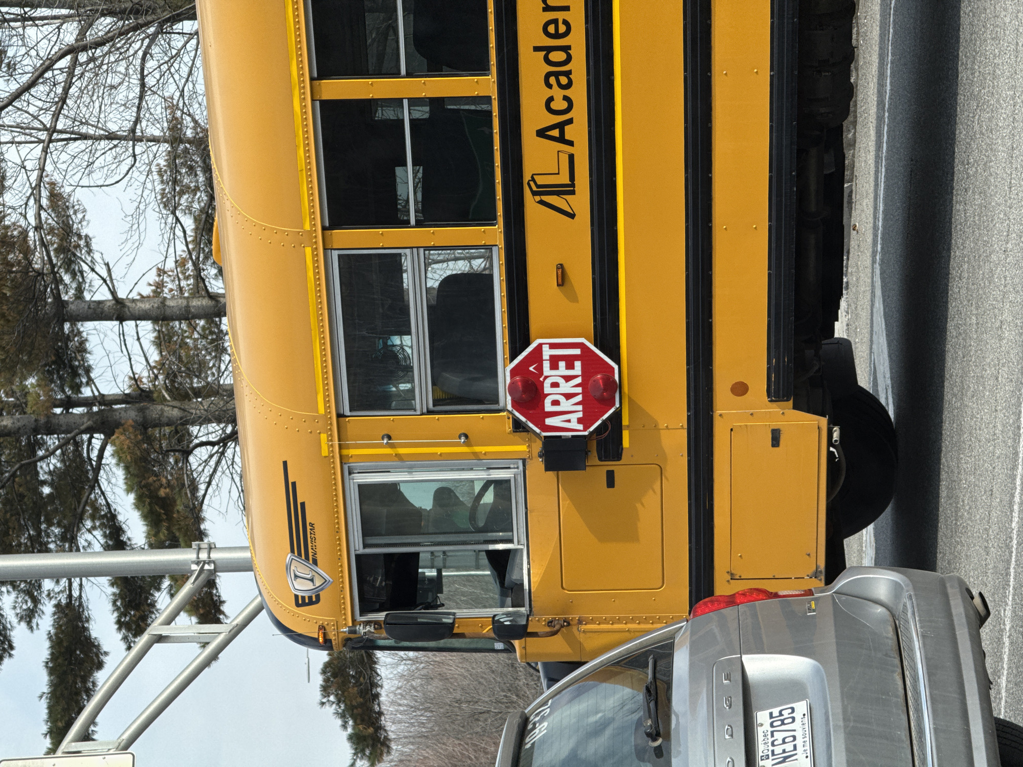 The image shows a yellow school bus with the word "Academie" on its side. The bus has its "ARRET" (STOP in French) sign extended. A silver Dodge vehicle is partially visible behind the bus, with the Quebec license plate "NE6785". The scene appears to be on a road in a residential area, with trees visible in the background. The location is likely Blainville, Canada, given the Quebec license plate.