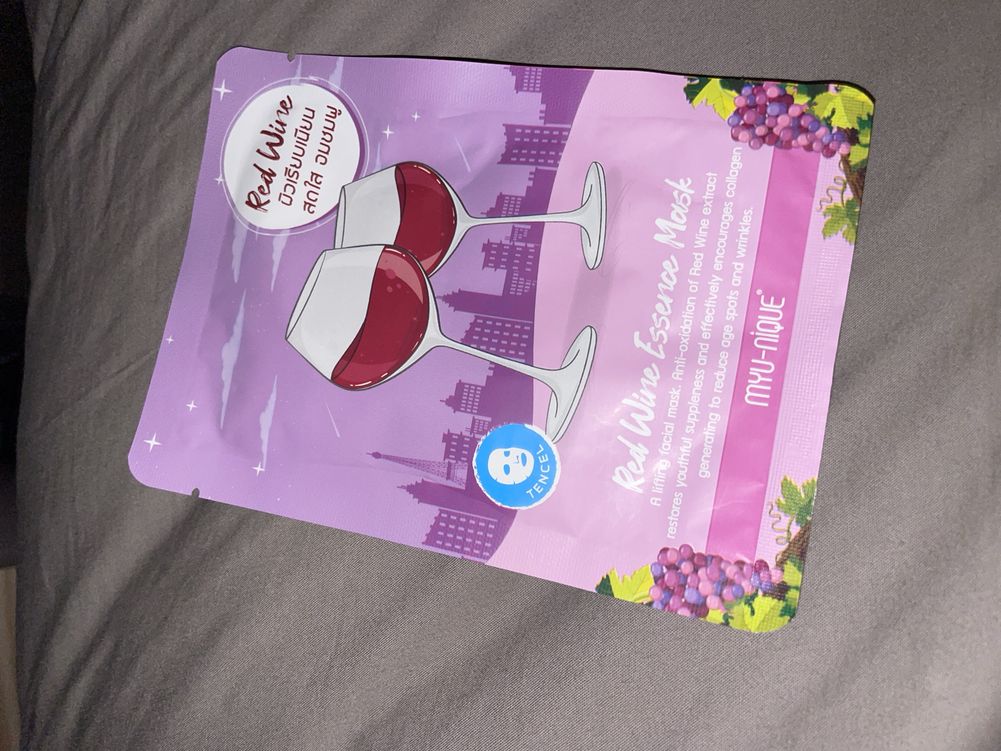The image shows a face mask package with a "Red Wine" theme. The package is purple and features illustrations of wine glasses, grapes, and a city skyline that resembles Paris, with the Eiffel Tower visible. There is text in English and what appears to be Thai. The brand name "MYU-NIQUE" is also visible. The mask package is lying on a grey, textured surface, likely a bed or couch. There is no discernible connection to the city of Umm Al Quwain, United Arab Emirates, in the image.