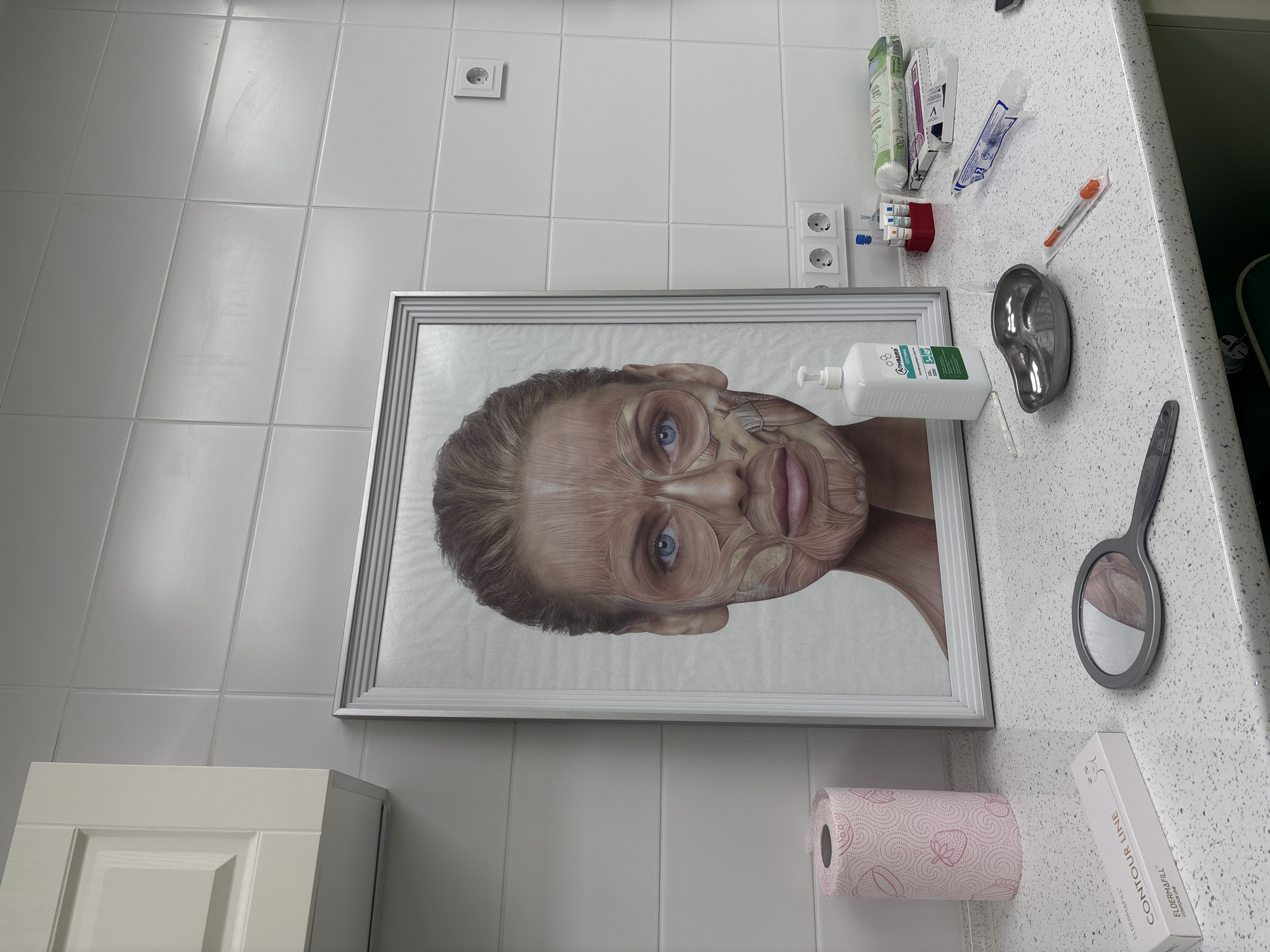 The image shows a room with white tiled walls and a white countertop. A framed picture of a human face with anatomical muscle details hangs on the wall. On the countertop, there are medical supplies, including syringes, needles, a small metal tray, and a bottle of antiseptic. Also present are a small hand mirror, a roll of pink toilet paper, and a box labeled "ELDERMAFILL CONTOUR LINE." The environment appears to be a medical or cosmetic treatment room. The location is not discernible from the image.