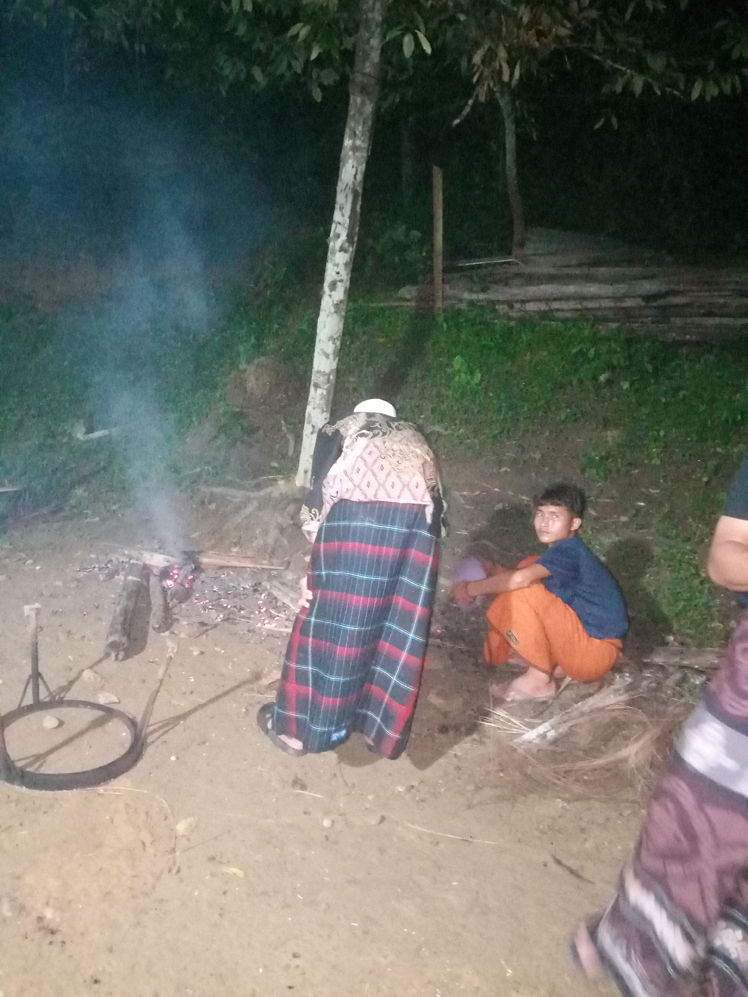 The image shows a nighttime outdoor scene in Lhokseumawe, Indonesia. In the foreground, a fire is burning with smoke rising. There are several people present. One person, viewed from behind, wears a patterned top and a plaid sarong, standing near the fire. Another person, a young male, sits on the ground to the right, wearing a blue t-shirt and orange pants, looking towards the camera. Part of another person's sarong is visible on the far right. The environment appears to be a natural setting with trees and foliage in the background. There are various objects scattered on the ground, including wood, ash from the fire, and a metal ring with support structures. The ground is rough and sandy. The lighting suggests artificial light sources are present, illuminating the scene despite the darkness.