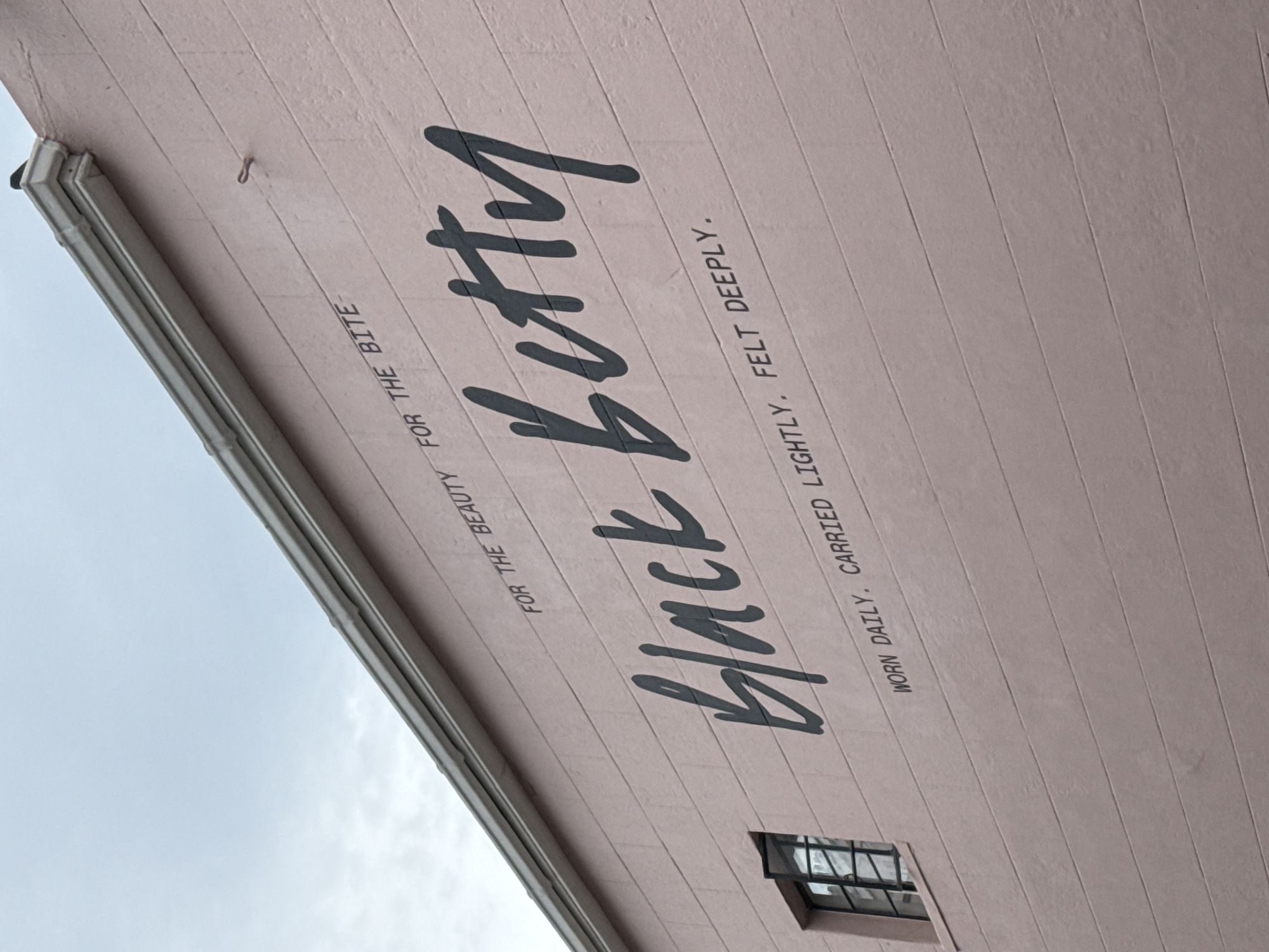 The image captures the exterior of a building, likely located in Cape Town, South Africa. The building is painted a light pink color, with the words "Black Betty" prominently displayed in a stylized font, along with smaller text "FOR THE BEAUTY FOR THE BITE," and "WORN DAILY. CARRIED LIGHTLY, FELT DEEPLY." The building's architecture features horizontal lines, a gutter system, and a small, partially opened window. The sky is overcast, indicating daylight conditions.