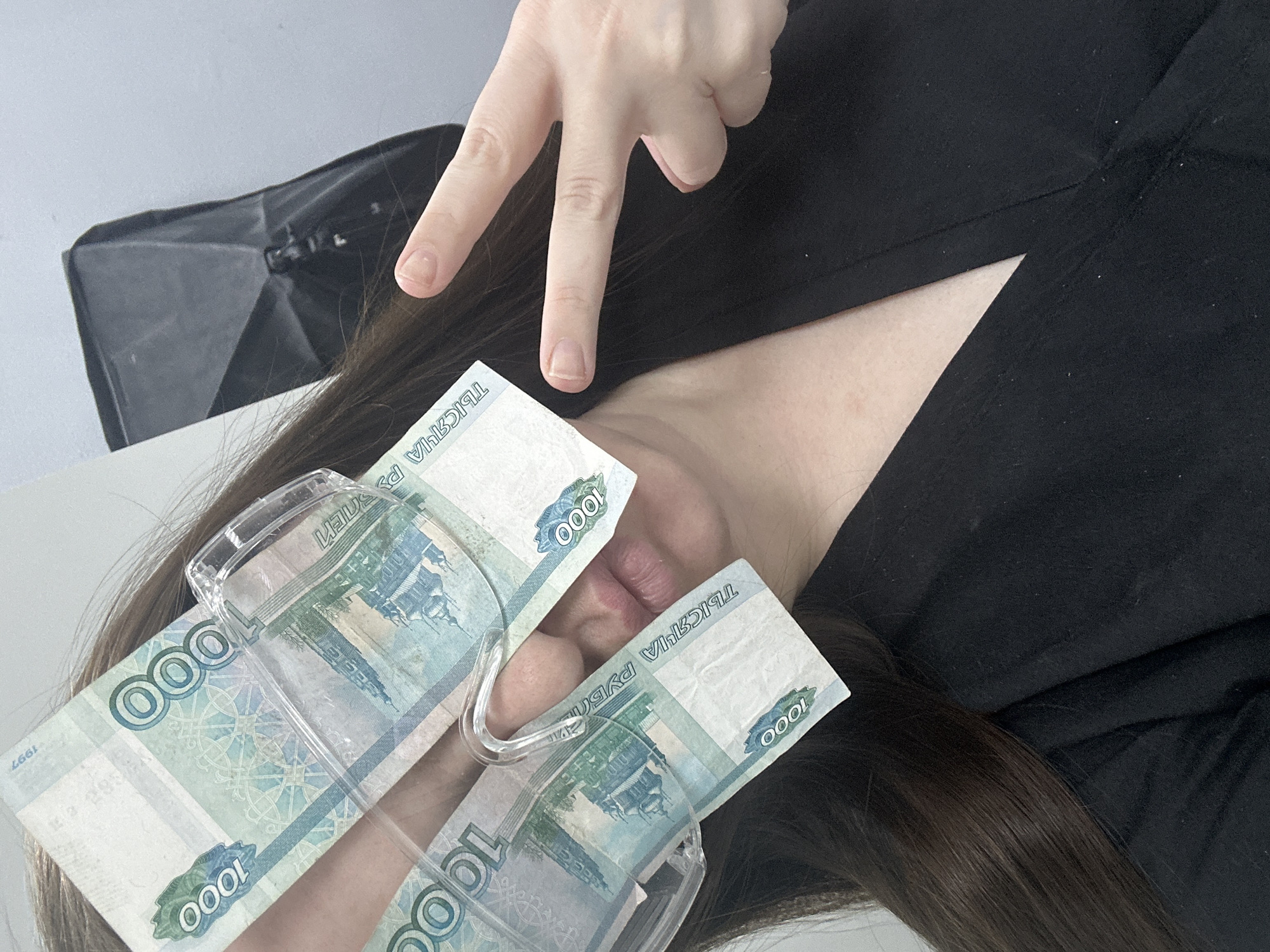 A close-up, high-angle photograph shows a person's face partially obscured by two 1000 Russian Ruble banknotes held in place by a clear plastic frame, resembling makeshift glasses. The person's lips are visible between the banknotes. One hand is raised, displaying a peace sign. The person is wearing a black garment. Long brown hair is visible. The background is a mix of white and gray surfaces. The location is identified as Chaikovsky, Russia based on the Russian currency.