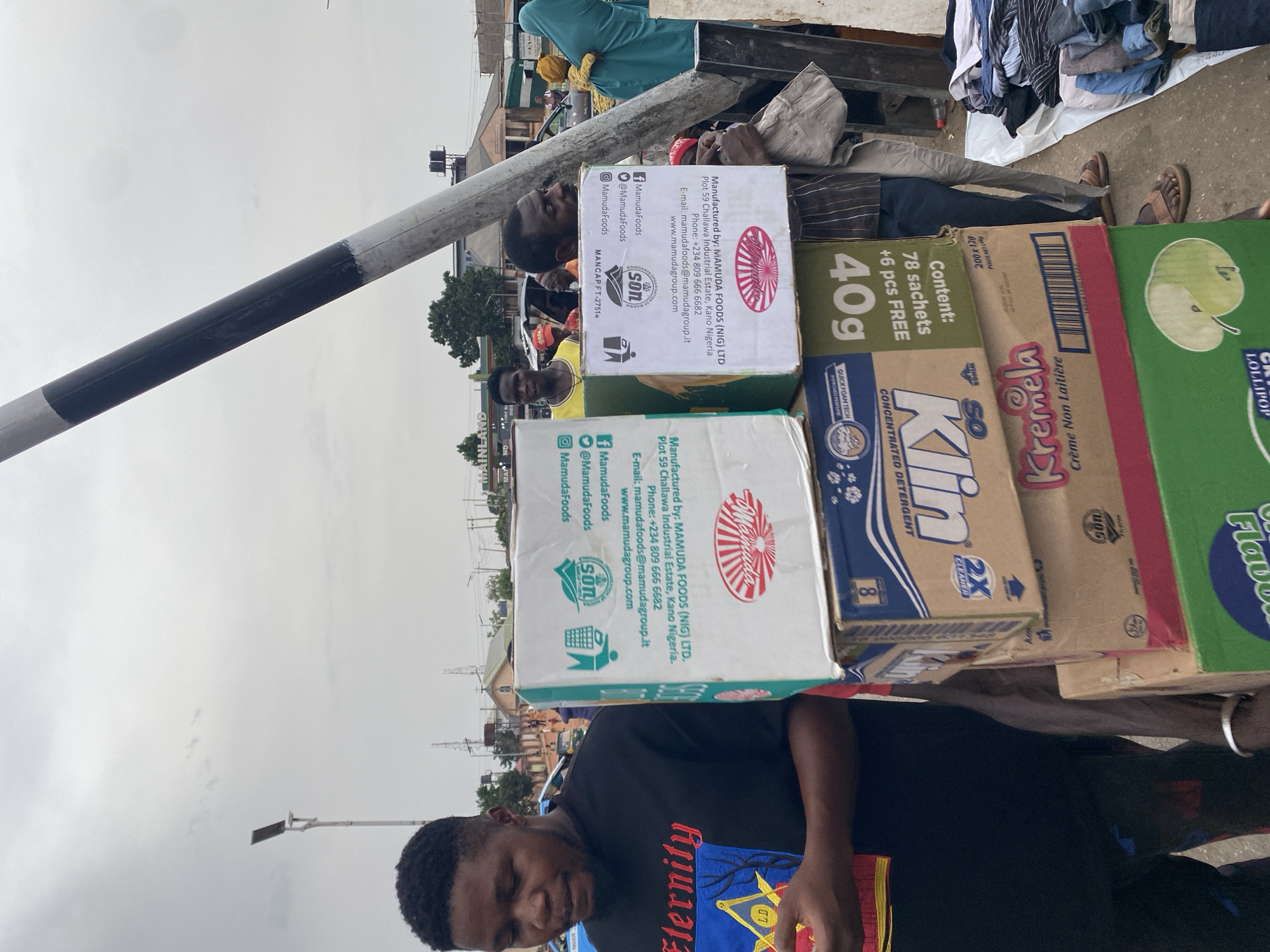 The image depicts a street scene in Gura Top, Nigeria. A man is carrying a stack of boxes on his head. The boxes are branded with the logo of Mamuda Foods (NIG) LTD, a company located in Kano, Nigeria. The boxes contain food products such as Klin detergent, Kremela Creme Non Laitiere, and GRR Lollipop. Other people can be seen in the background, along with a variety of objects, including clothes and a black and white striped pole. The sky is overcast, and the overall environment appears to be a busy urban area.