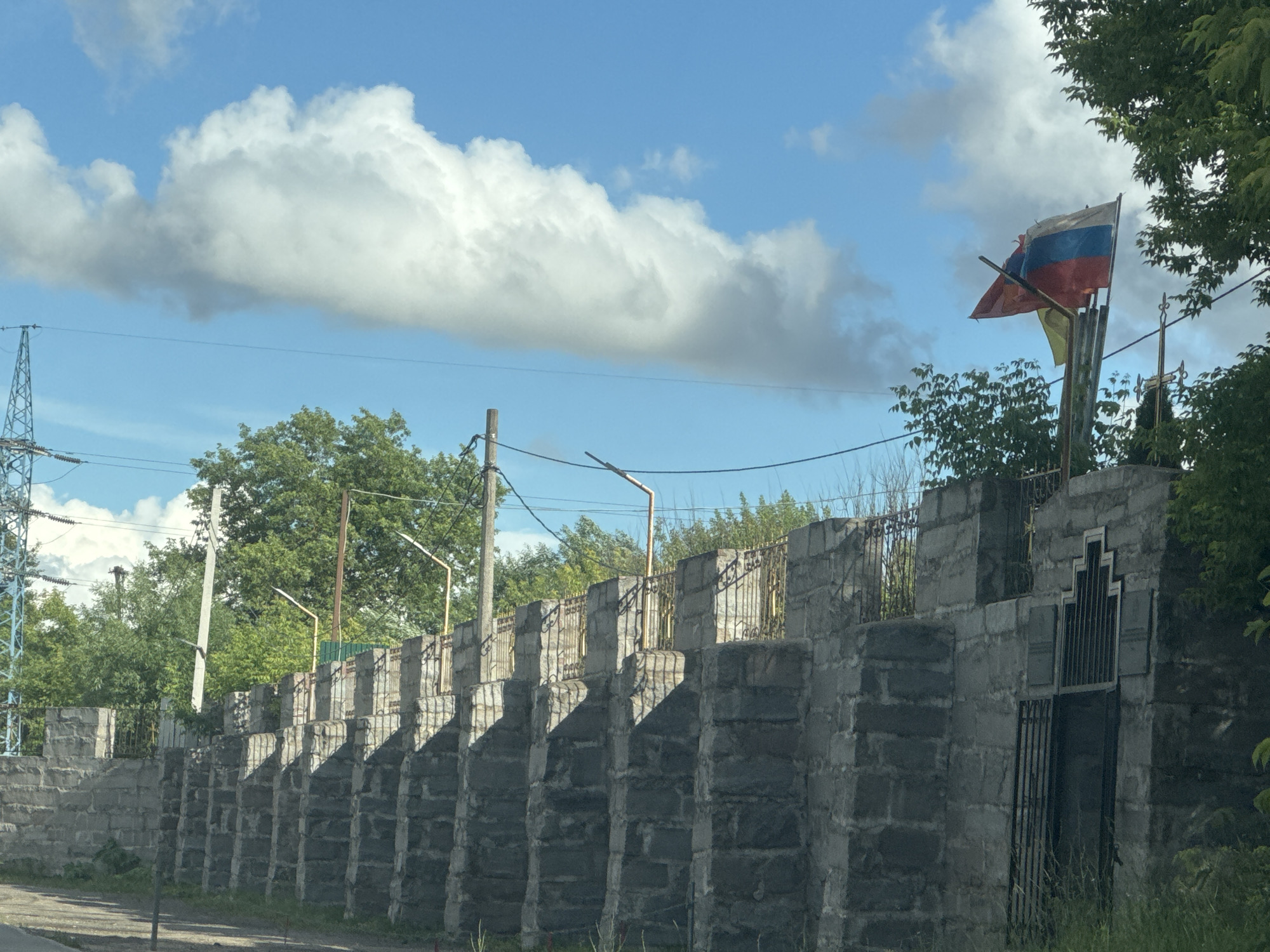 The image shows a view of a stone wall with a Russian flag flying above it. The wall is constructed from large, grey stones and has a series of evenly spaced vertical supports topped with decorative metalwork. Behind the wall, trees and vegetation are visible, along with utility poles and wires. The sky is blue with large, white clouds. The Russian flag, with its horizontal white, blue, and red stripes, is prominently displayed on a flagpole, appearing to be slightly worn. A yellow flag is also visible behind it. A metal gate is set into the wall on the right side of the image. The overall scene suggests an outdoor setting on a sunny day, likely in Russia given the flag. There is no visible text in the image. The image appears to be taken from a low angle, possibly from a vehicle.