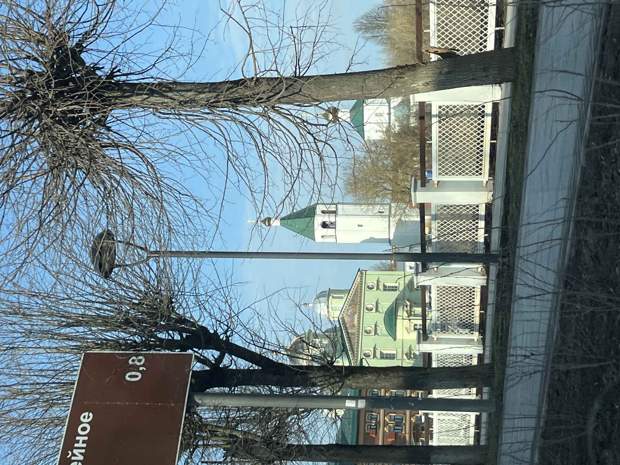 The image shows a street in Klin, Russia, with a view of several buildings, likely religious structures, through bare trees and a white fence. A street sign with Russian text and the number "0,8" is visible in the foreground. The buildings in the background have distinct architectural features, including green roofs, domes, and a bell tower. The sky is blue. The image appears to be taken from inside a vehicle due to reflections on the glass.