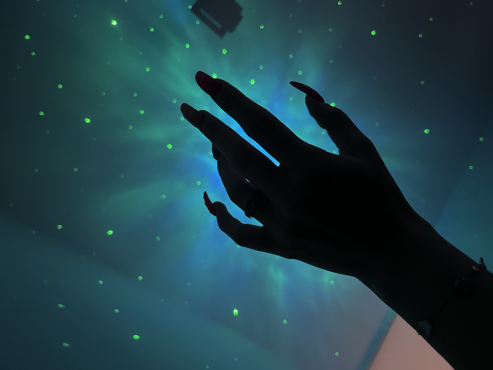 A low-angle shot depicts a person's hand with long, red fingernails silhouetted against a blurry background of blue and green lights that resemble stars. The hand is outstretched as if reaching or interacting with the light. The scene likely takes place indoors, possibly in a darkened room illuminated by a projector or decorative lighting. A ring is visible on one of the fingers. A bracelet is visible on the wrist. There is no information available to confirm this picture was taken in Plovdiv, Bulgaria.