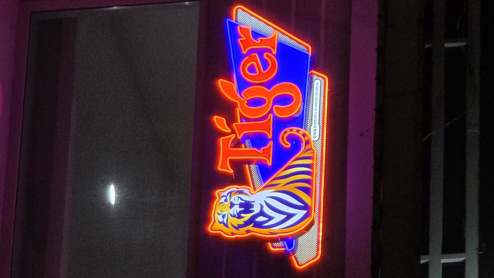 The image shows a vertical neon sign for "Tiger," a beer brand, mounted on a dark surface. The sign features a stylized tiger illustration in blue, yellow, and white, with the word "Tiger" vertically stacked above it in red neon. The sign is located in Nha Trang, Vietnam, as indicated by the Vietnamese text visible on the sign's edge. To the left of the sign is a doorway or window, and to the right, a metal structure or gate. The overall lighting suggests it is nighttime.