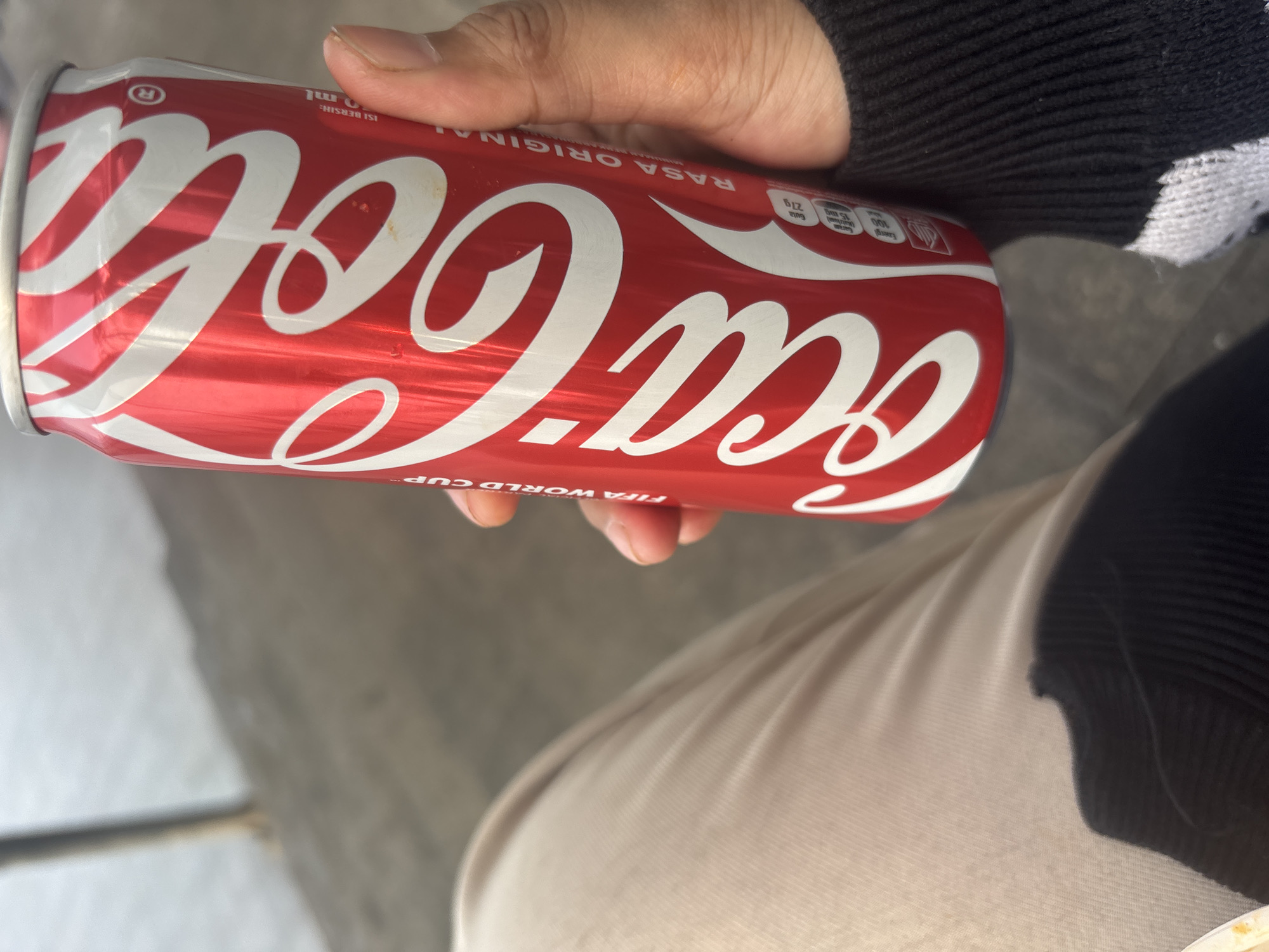A person's hand is holding a Coca-Cola can. The can is red with white lettering. The background is blurred and appears to be outdoors, with a gray, concrete surface and some greenery. The person is wearing a black sweater with white accents. The image is taken from a close-up perspective, focusing on the can and the hand. There is no indication of the specific location within North Jakarta, Indonesia.