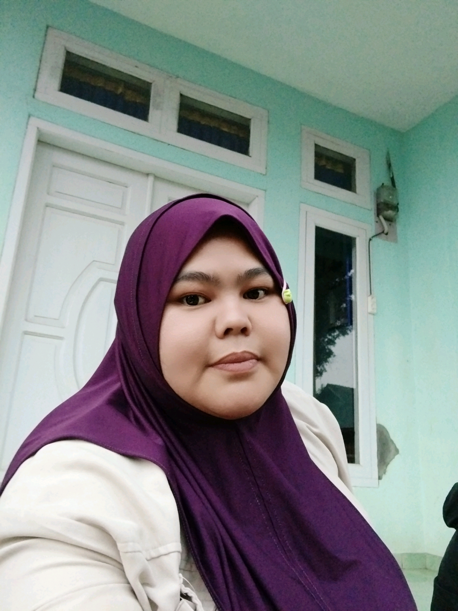 A portrait photograph shows a young woman wearing a purple hijab and a light-colored jacket. She is looking directly at the camera with a neutral expression. The background consists of a light blue wall with white trim around a door and two windows. A green electrical box is mounted on the wall to the right of the windows. The image appears to be taken outdoors.