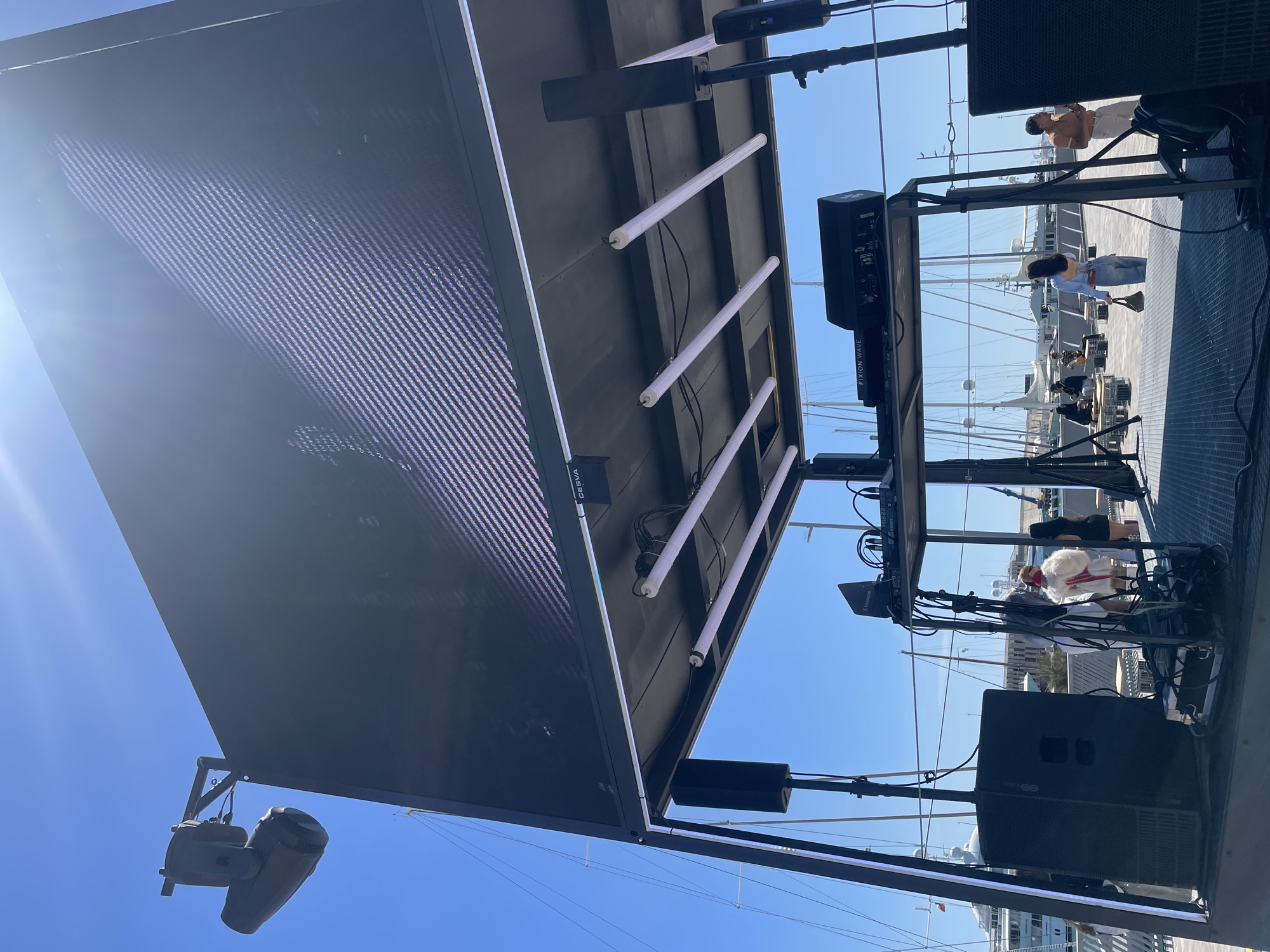 This media file captures an outdoor stage setup against a clear blue sky, likely in a marina or port area like Barcelona, Spain. The image is taken from a low angle, looking up at the underside of the structure and the elevated platform.
The primary subjects are a large, rectangular LED screen, visible on the left, displaying a pixelated, striped pattern. Mounted beneath a canopy or platform are three prominent, long tubular white lights, possibly LED bars. Various professional audio and lighting equipment are integrated into the stage design. Two large black speakers are visible, one explicitly labeled "FIXXION WAVE" and another "HK AUDIO". Below the screen, two moving head lights are mounted.
On an elevated platform to the right, several individuals are present. One person in a light top and pants stands prominently, while another in a light blue top and jeans appears to be holding an object. Further back, more people are visible, including one in a white, fluffy garment. On this platform, a mixing console or similar audio control unit is set up, with a "SENNHEISER" branded component attached. Cables are extensively visible, connecting the different pieces of equipment.
The scene suggests an active outdoor event, such as a concert, public viewing, or performance. The presence of performers/crew on the platform, along with the sophisticated technical equipment, indicates preparation for or the midst of a live show. The clear blue sky and bright sunlight, accompanied by a lens flare on the upper left, denote a sunny day, likely during morning or early afternoon hours. The background features several sailing yachts and boats, confirming a waterfront location. Other visible text includes "GESVA" or "CESVA" partially seen near the LED screen.