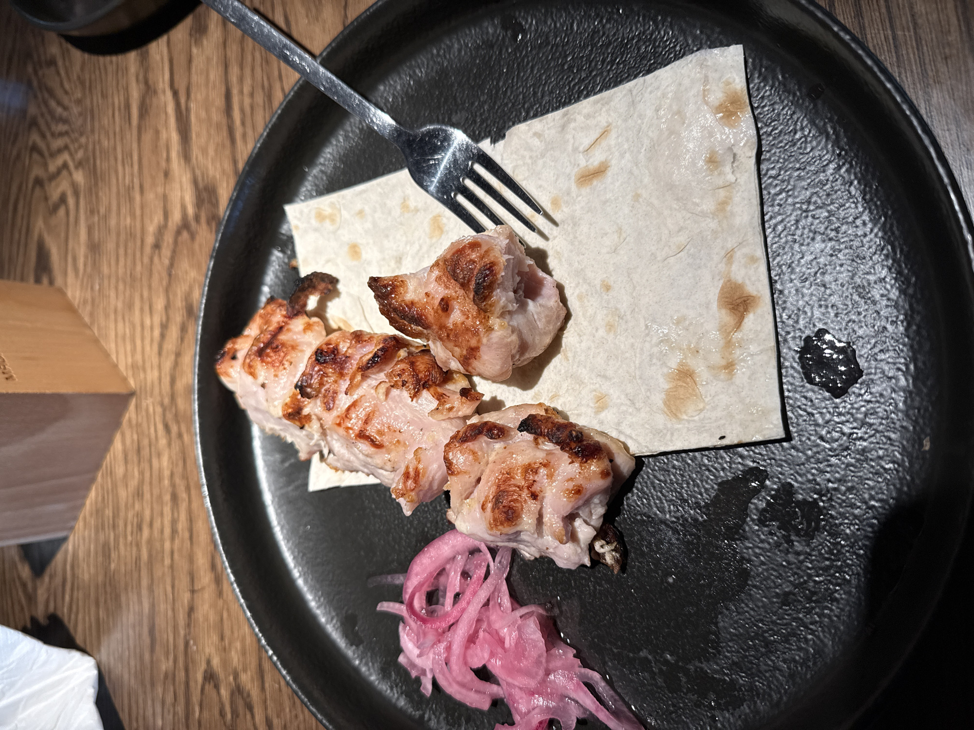 The image shows a close-up, overhead view of a dark plate on a wooden table. On the plate are pieces of grilled meat, likely chicken or pork, served on a piece of flatbread. A silver fork rests on the flatbread, with its tines pointed towards the meat. To the side of the meat is a small pile of thinly sliced pickled red onions. There are some oily splotches on the flatbread and the plate. In the background, a portion of a wooden object, possibly a table leg or a decorative element, is visible. The lighting suggests an indoor setting, possibly a restaurant, with artificial light. The overall presentation suggests a meal is being served or has just been served. There is no visible text in the image.