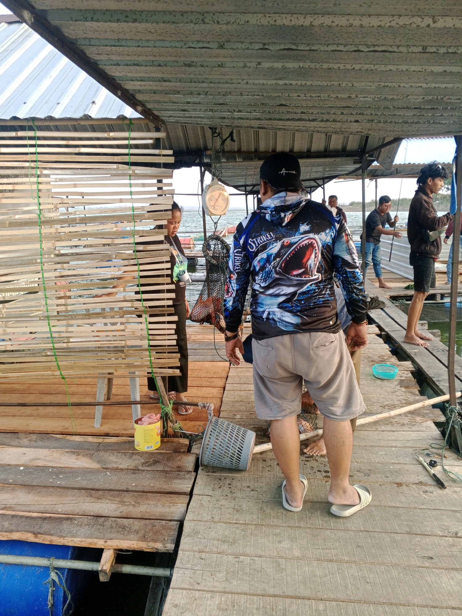 The image depicts a group of people on a wooden platform over water. The platform appears to be a fishing area, with fishing nets and a scale visible. Several individuals are present, some engaged in fishing activities and others observing. The architecture suggests a rustic, possibly temporary structure, with a corrugated metal roof supported by wooden beams. The background shows a body of water with a distant shoreline. The people are dressed in casual attire, with some wearing fishing-themed clothing. The overall scene conveys a sense of everyday life and recreational activity in a waterside community. The location is identified as Sendang, Indonesia.