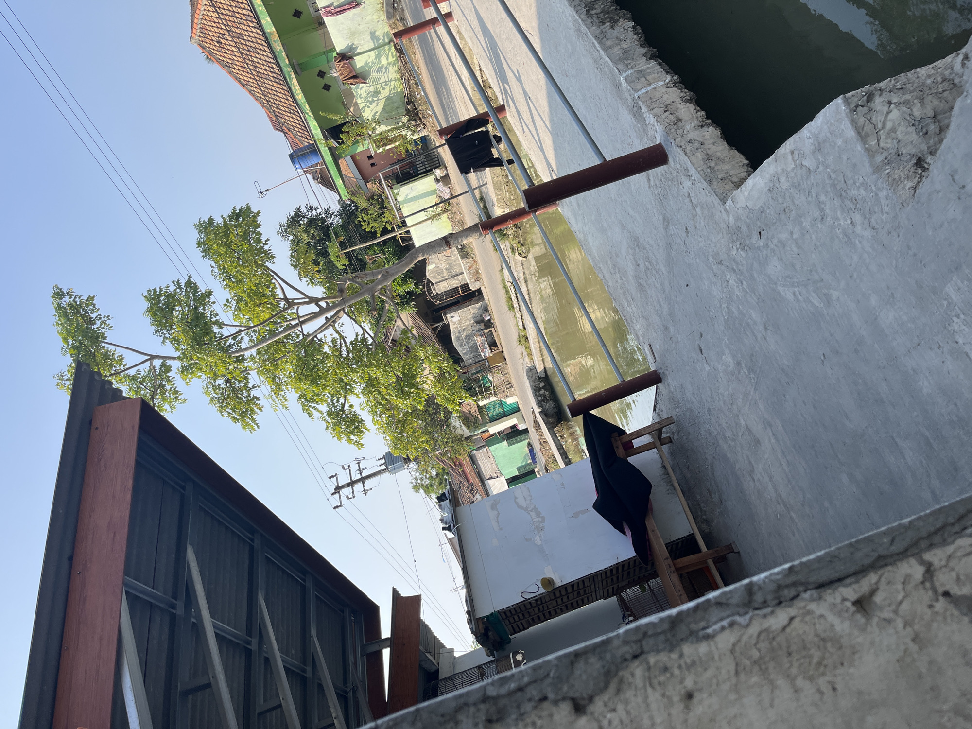 This image depicts a canal-side view in Laban, Indonesia, likely on a sunny day given the clear blue sky and sharp shadows. In the foreground, a concrete walkway runs alongside a murky canal. A simple wooden bench with a dark cloth draped over it is visible to the left. Beyond the canal, a street is lined with houses. One house in the mid-ground is painted green with a red-tiled roof. A tree with vibrant green leaves stands prominently in the center of the frame, its branches reaching towards the sky. Electrical poles and wires crisscross the upper portion of the image, indicating the presence of infrastructure. A black jacket is hanging from a railing along the canal. The overall scene suggests a residential area adjacent to a water channel, common in many parts of Indonesia.
