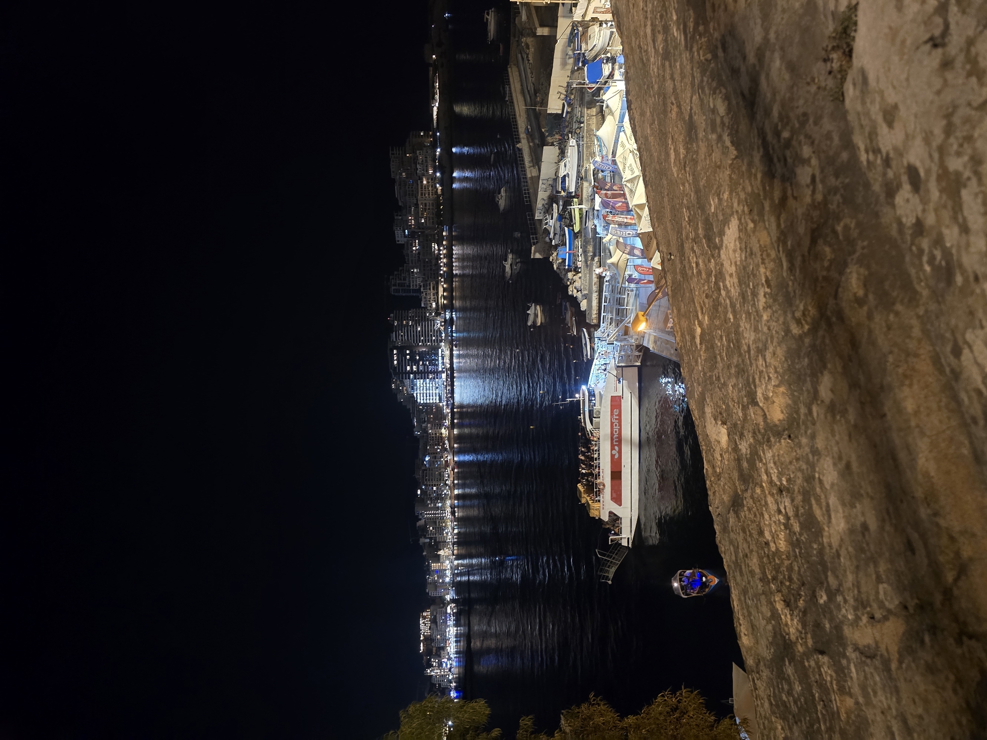 This is a night-time, low-angle, vertical shot of Valletta's Grand Harbour, taken from the Upper Barrakka Gardens. The foreground is dominated by a rough-hewn, stone wall, likely part of the fortification. To the left, a dark, leafy tree branch partially obscures the view. The harbor is illuminated by the lights of the city, reflecting on the water.
In the mid-ground, a large ferry, branded with "mapfre," is docked, carrying numerous passengers visible on its upper deck. A smaller, illuminated boat with several occupants is navigating the water in front of the ferry. Further into the harbor, various other boats and smaller vessels are visible, some docked and others on the water.
The background showcases the illuminated cityscape of Valletta, with rows of buildings displaying numerous lit windows. The skyline is distinct, characterized by modern and older architectural styles. The overall scene depicts a vibrant night in a coastal city, with activity on the water and the urban landscape aglow.