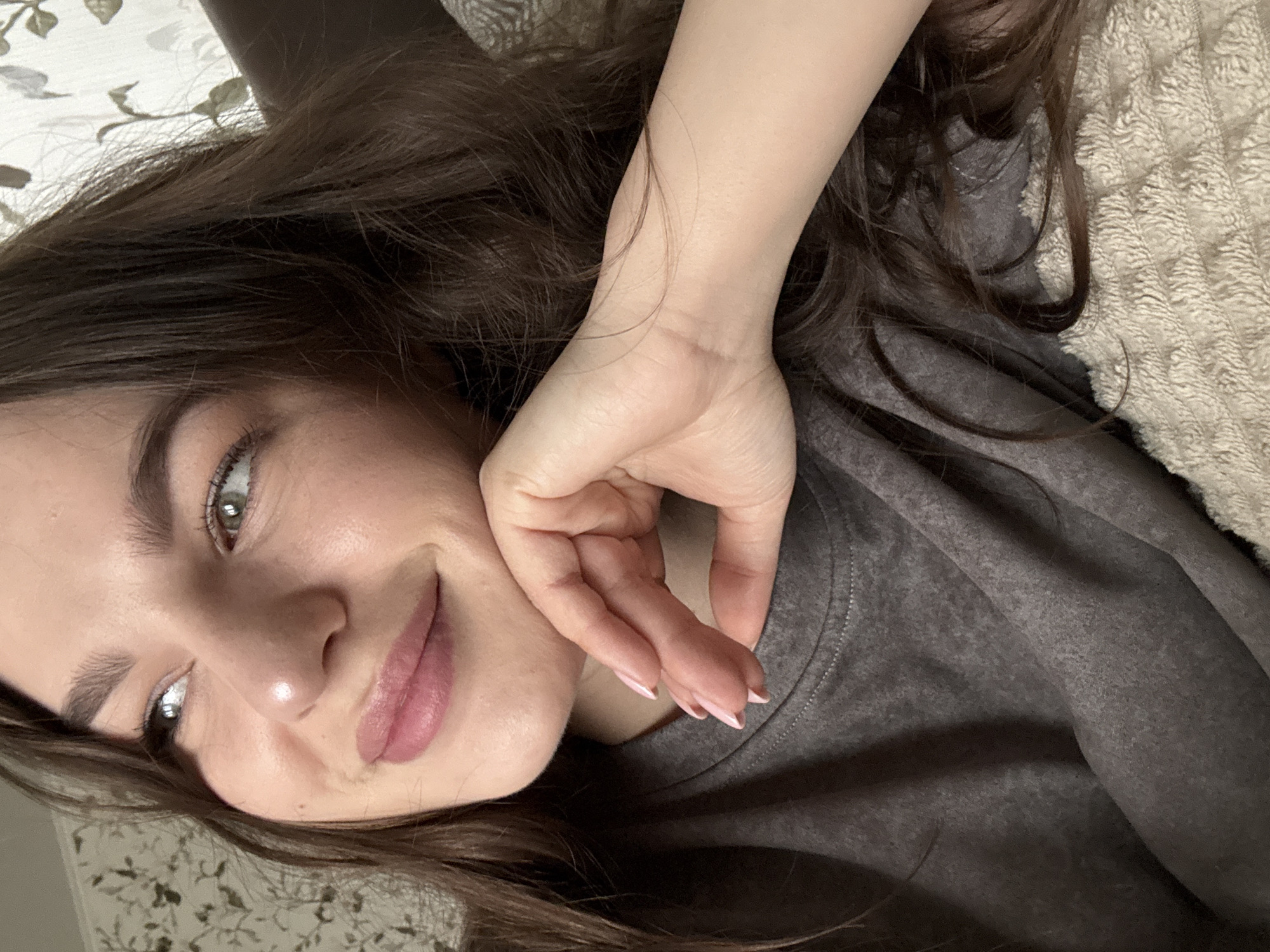The image shows a woman indoors, lying down and holding her hand near her face. She has long, dark brown hair, fair skin, and is wearing a gray shirt. She has light eye makeup and pink lipstick. The background includes a textured blanket or bedspread. There is no visible information in the image to confirm the location is in the city of Vnukovo, Russia.