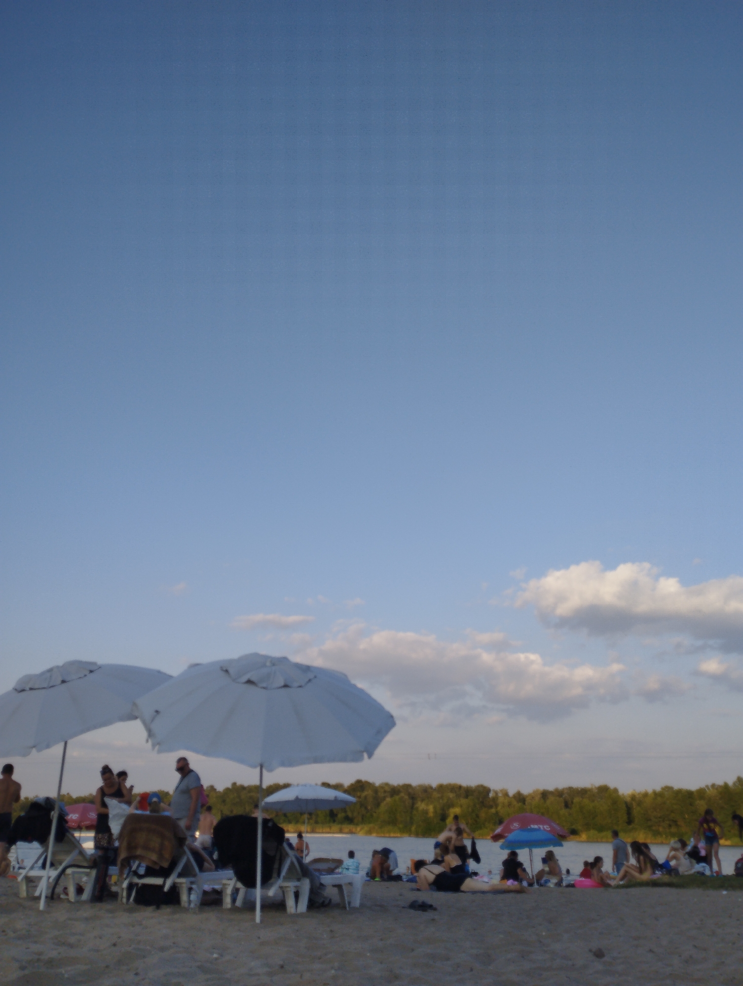 The image depicts a crowded beach scene on a sunny day, with many people relaxing under umbrellas and on lounge chairs. The sandy beach is filled with beachgoers, some of whom are sunbathing, while others are socializing or enjoying the water. The background shows a lush green forest bordering a body of water, likely a lake or river, under a clear blue sky with scattered white clouds. The overall atmosphere suggests a recreational gathering, possibly a public beach or a resort area, where people are enjoying leisure activities. The image's composition is wide, showcasing the breadth of the beach and the density of the crowd.