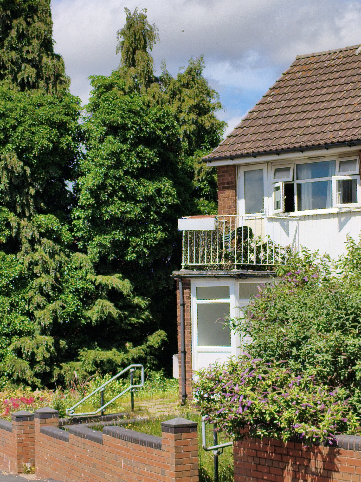 The image depicts a residential building with a balcony and a brick wall in the foreground, set against a backdrop of lush green trees under a partly cloudy sky. The building appears to be a typical terraced house or apartment block, with white-painted walls, brown tiled roof, and a white balcony railing. One window on the balcony is open. In the foreground, a low brick wall with stone capping runs across the image. A metal handrail ascends from a lower level to the right of the wall. Dense green foliage, including flowering bushes with small purple blossoms, borders the lower part of the building and the wall. The scene suggests a suburban or urban residential area, likely in Sandwell, United Kingdom, given the architectural style and vegetation.