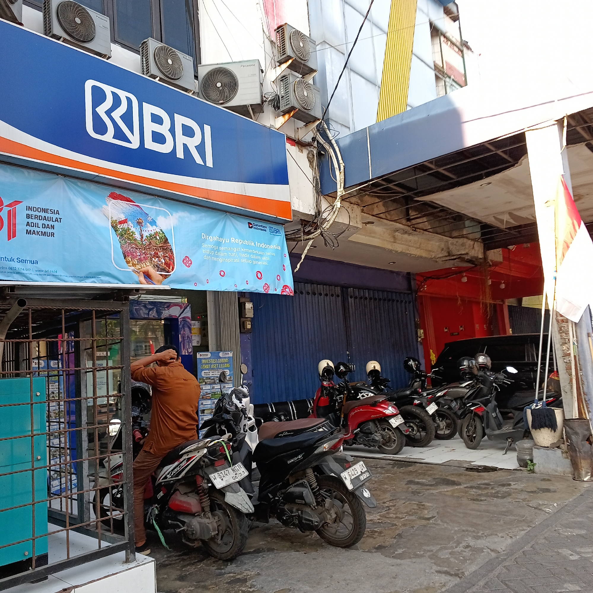 The image shows the exterior of a BRI (Bank Rakyat Indonesia) branch in Wedoro, Indonesia. A large blue sign with the BRI logo and name is prominently displayed on the building. Below the sign, a light blue banner features Indonesian text, including "Dirgahayu Republik Indonesia" and the BRI logo. Air conditioning units are mounted on the building's exterior.

In front of the bank, several motorcycles are parked. One person, wearing a brown shirt and pants, is seated on a motorcycle, with their hand touching their head. Another motorcycle in the foreground has a license plate that reads "W 3147 NGR". Further back, other motorcycles are visible, including a red one and several black ones. The ground is paved with concrete. The scene appears to be a street-level view of a commercial area.