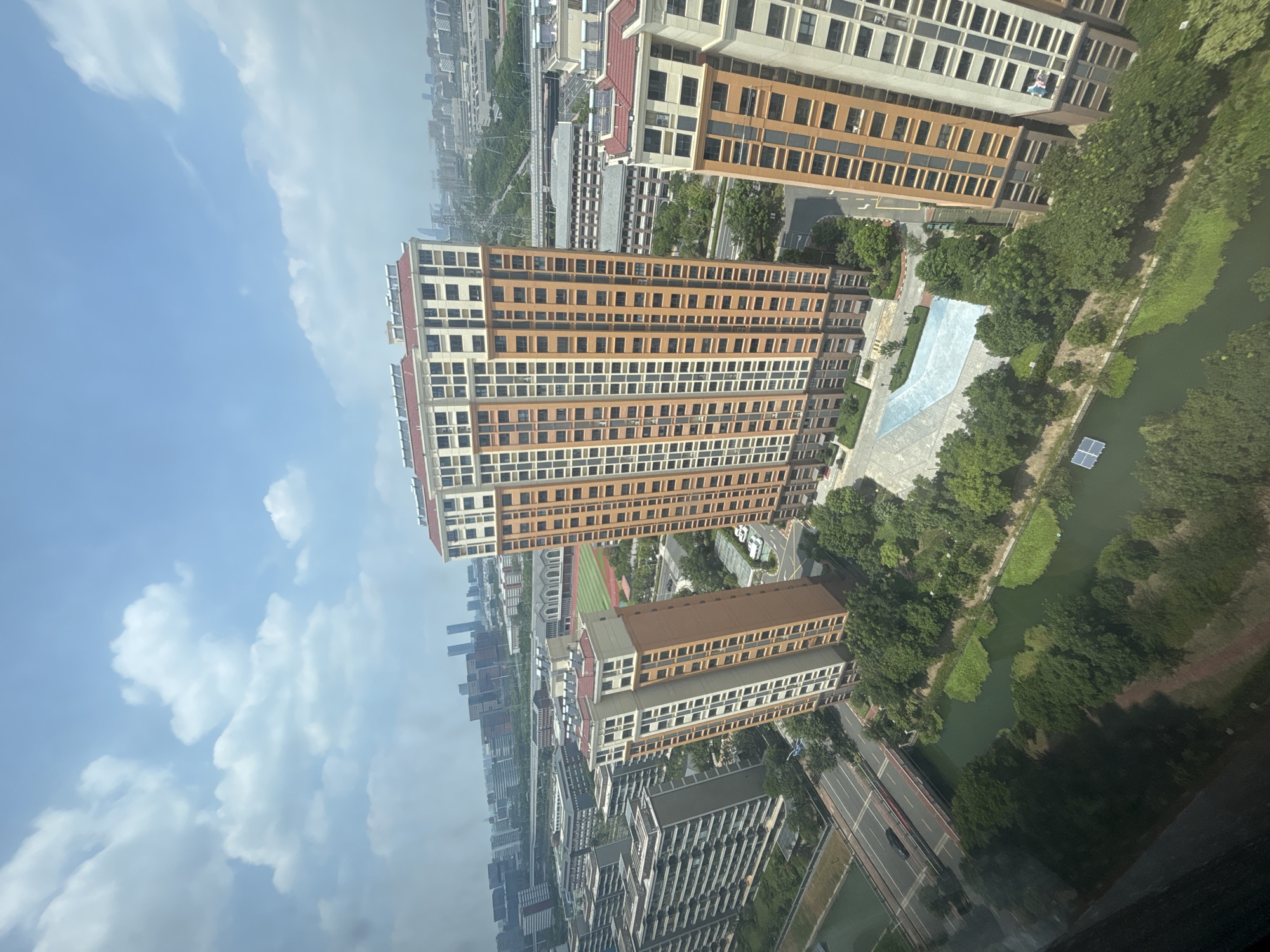 The aerial, high-angle view captures a modern urban landscape in Suzhou Industrial Park, China. The scene is dominated by several tall, residential apartment buildings with distinctive orange and cream facades, some featuring red-tiled roofs. In the foreground, a green, algae-filled canal winds through the cityscape, with a road and a small bridge crossing it. Lush green trees line the canal and surround the buildings. A paved recreational area with a light blue surface is visible near the base of the buildings. Further in the background, more residential blocks, administrative buildings, and a stadium with a red track are scattered across the urban expanse under a bright blue sky with scattered white clouds. The overall impression is of a densely populated, well-developed urban area.