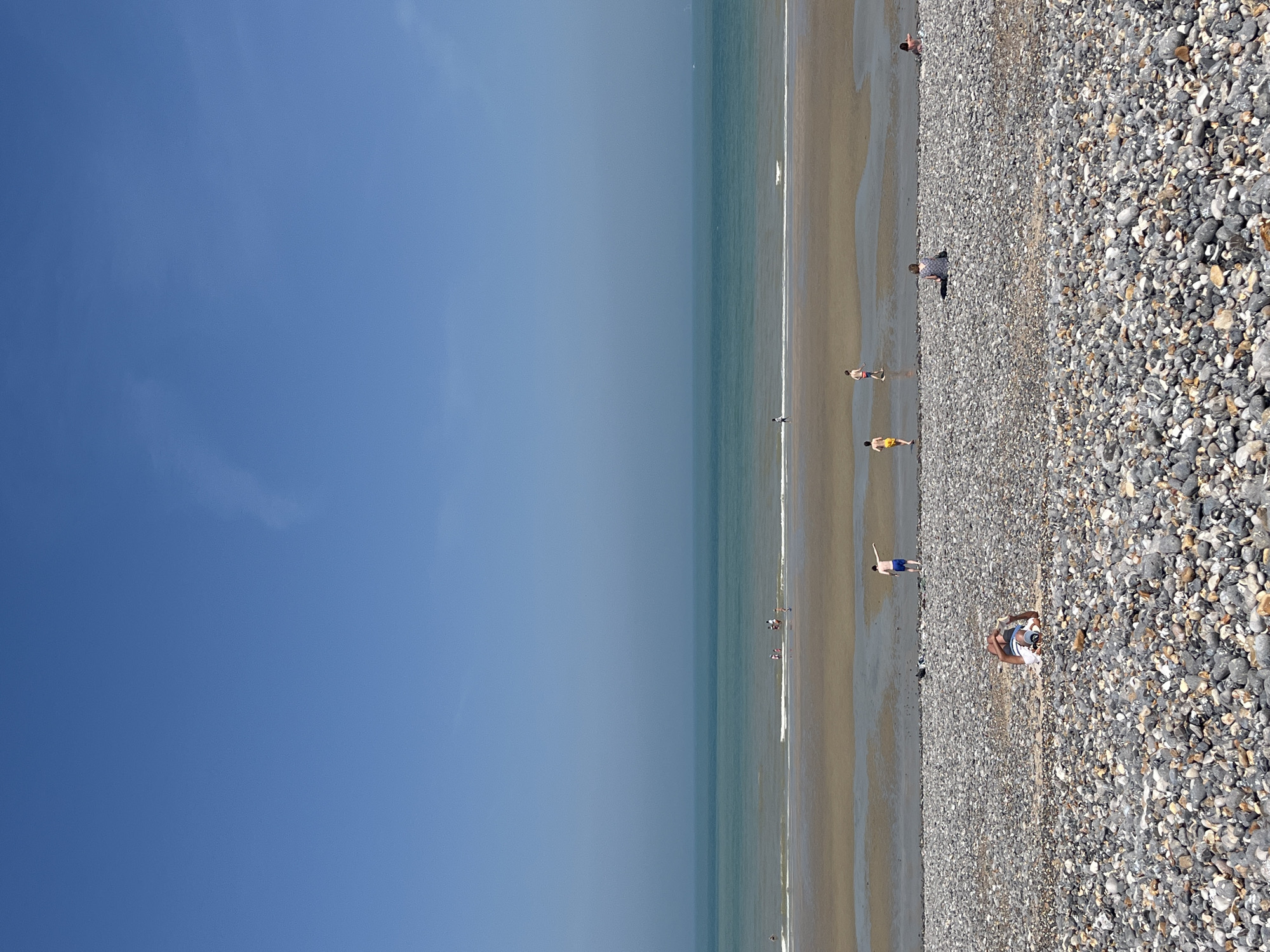 The image captures a view of the pebble beach and the English Channel in Dieppe, France. The sky is a clear, bright blue with a faint haze on the horizon. The sea is a tranquil turquoise, with small waves gently lapping at the shore.
The foreground is dominated by a vast expanse of pebbles, ranging in color from white and grey to darker hues. Several people are scattered across this pebble beach, either relaxing or sunbathing. In the middle ground, the wet sand of the receding tide creates reflective pools. A few individuals are walking or playing in the shallow water. Further out, more people are visible near the water's edge. The overall impression is one of a peaceful, sunny day at the beach.