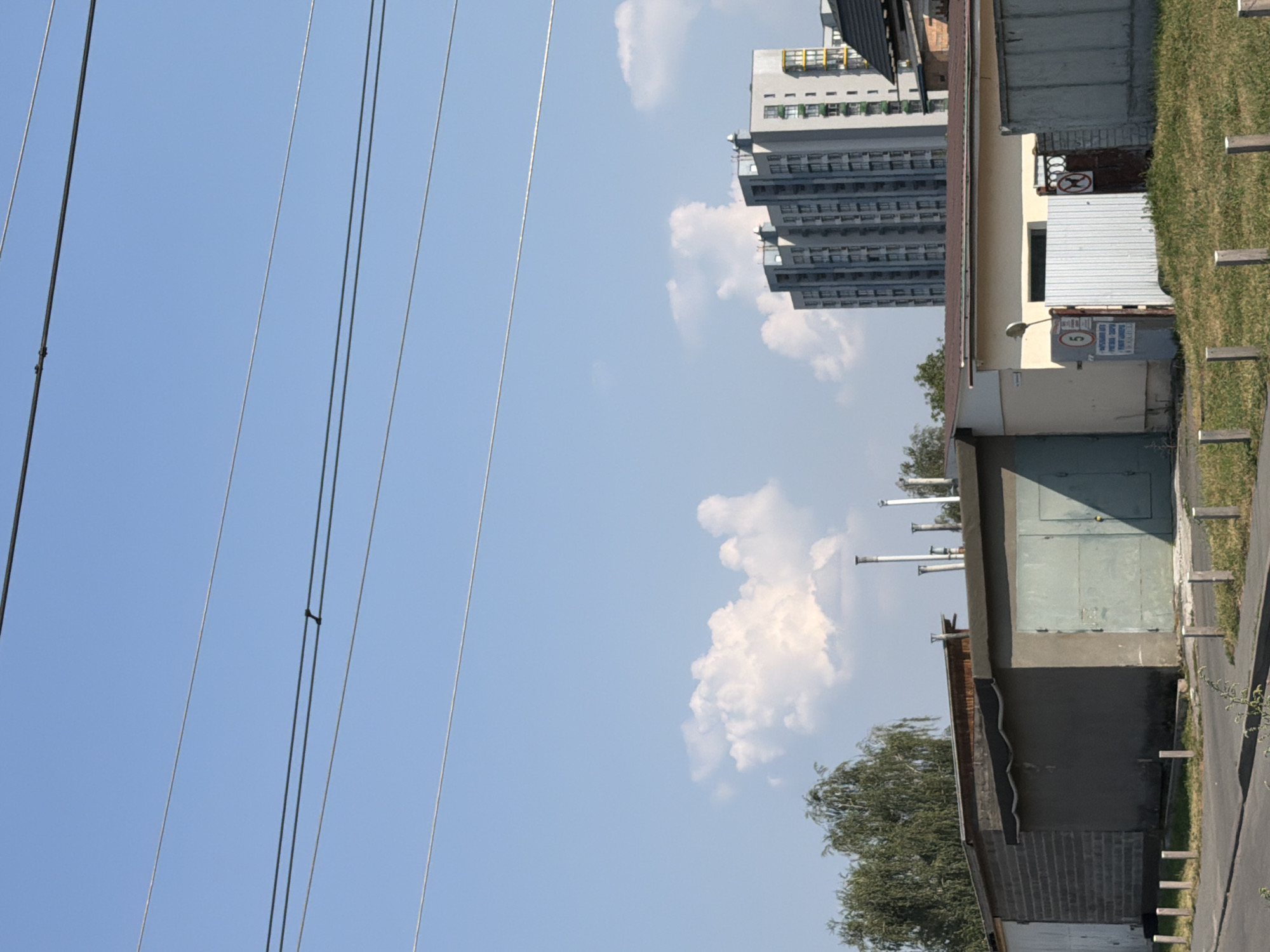 The image depicts a street scene in Kyiv, Ukraine, under a clear blue sky with scattered white clouds. In the foreground, a paved road runs horizontally across the bottom of the frame, bordered by a grassy area with wooden posts. Beyond the grass are several low-rise buildings that appear to be garages or storage units, constructed from brick and concrete. One garage has a light blue metal door. Further back, a modern, tall apartment building with a gray facade and numerous windows stands prominently. A tree with green foliage is visible to the left of the apartment building. Power lines are strung across the upper portion of the image. The overall impression is one of a residential or industrial area within the city.