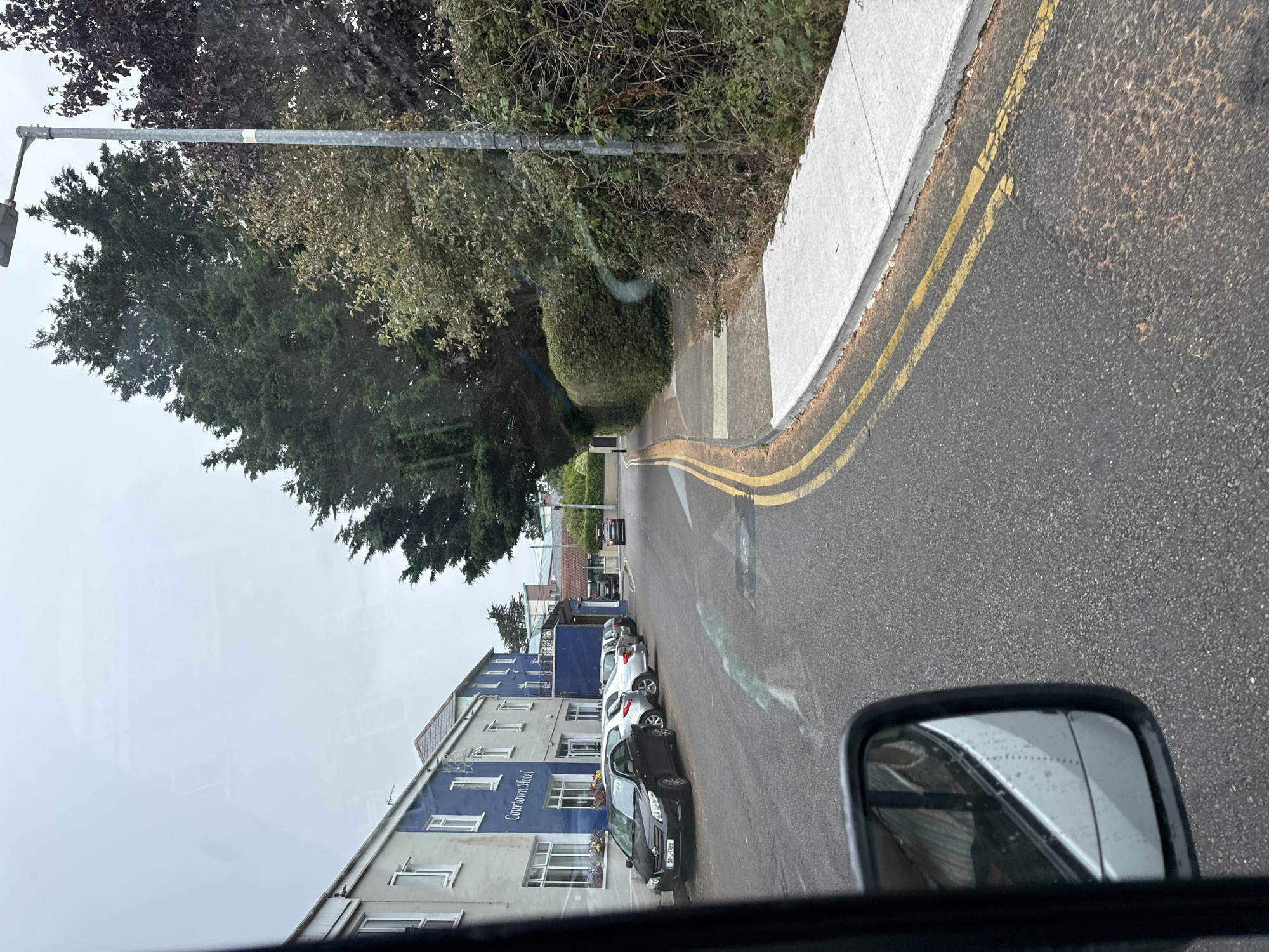 The image depicts a street scene in Courtown, Ireland. A blue and white building labeled "Courtown Hotel" is visible on the left side of the road, with several cars parked in front of it. The road itself is asphalt with double yellow lines marking the center. A sidewalk runs alongside the road, bordered by a hedge and trees. A streetlamp stands on the right side of the road, and a single black car is driving away in the distance. The weather appears overcast. The reflection in the side mirror of a vehicle suggests the photo was taken from inside a car.