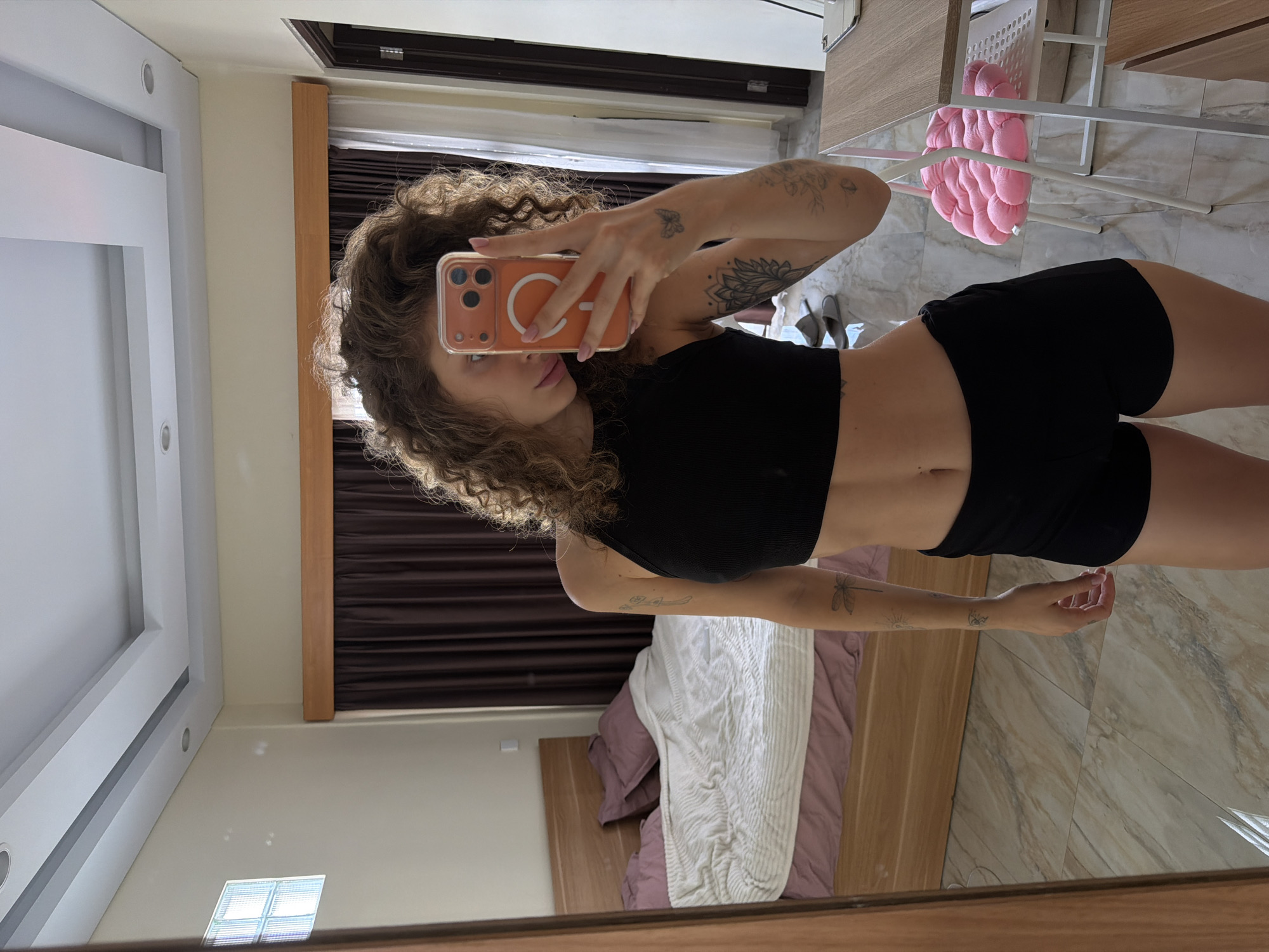 A mirror selfie captures a young woman in what appears to be a modern bedroom or hotel room, typical of North Kuta, Indonesia. The woman has curly brown hair and a slim physique, visible in her black ribbed crop top and black shorts. She is holding an orange smartphone, obscuring part of her face, with a white circular MagSafe ring visible on the phone case. Her fingernails are painted light pink.
Several tattoos adorn her arms: a small butterfly on her right bicep, a prominent mandala-like design on her left forearm, a dragonfly on her right forearm, and other smaller sun and wave motifs.
The background, seen in the mirror reflection, features a white bed frame dressed with a light purple duvet and a white textured blanket. Dark brown pleated curtains cover a window, framed by light brown wood. The room has a light-colored, possibly marble or ceramic, tiled floor and a white ceiling with recessed lighting. To the woman's right, a white metal rack or shelf holds a distinctive pink, textured, cloud-shaped cushion or pillow. A pair of grey slippers rests on the floor nearby. The bright, even lighting suggests it is daytime. No discernible text is visible on any items.