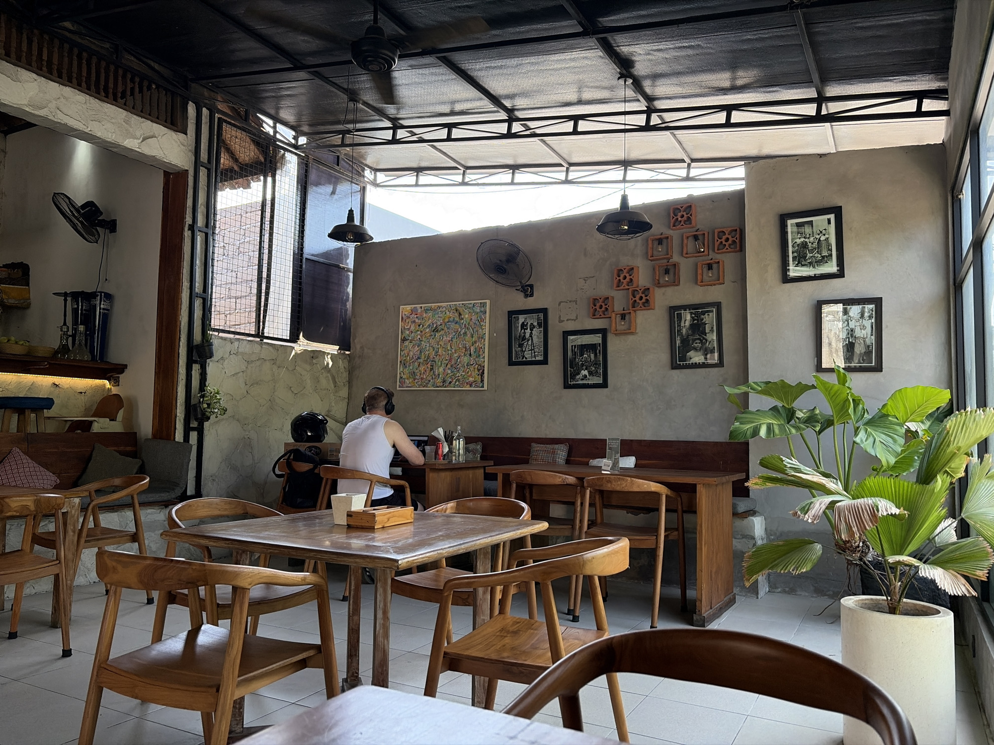 This image depicts the interior of a cafe, possibly a beachside establishment in Kubu, Indonesia, with an open-air feel. The space features a combination of concrete and natural materials. A person wearing a white tank top and headphones is seated at a wooden table, facing a laptop. The cafe is furnished with numerous wooden tables and chairs, some of which are occupied by patrons. The walls are adorned with framed black and white photographs and a colorful abstract painting. Large windows allow natural light to enter, and several ceiling fans are visible. A large potted plant with broad green leaves adds a touch of nature to the scene. The overall ambiance suggests a relaxed and casual dining environment.
