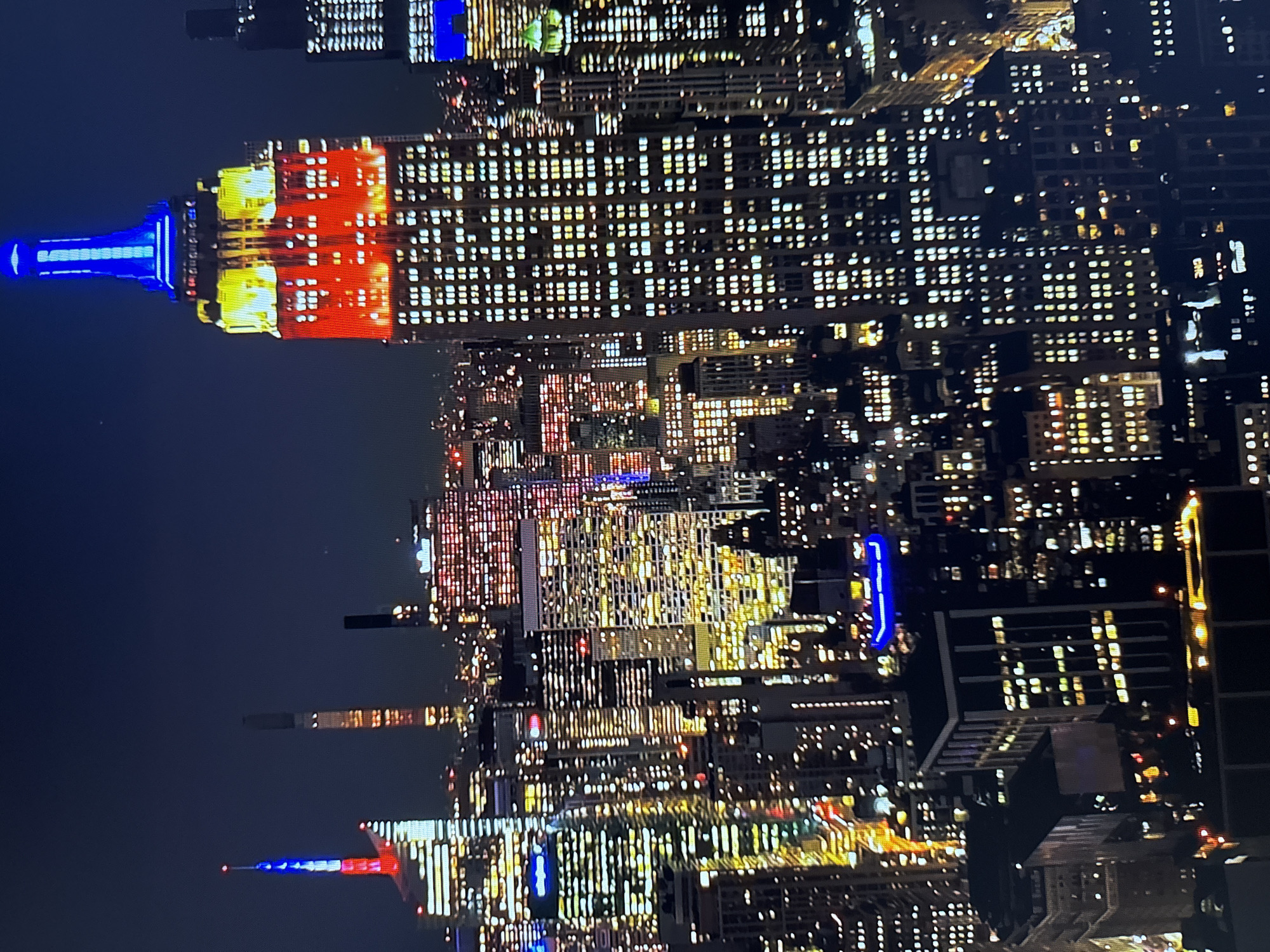 The image shows a nighttime aerial view of a densely packed cityscape, dominated by illuminated skyscrapers. The Empire State Building is prominently featured in the center-right, its spire illuminated in blue and yellow, with the main body of the building lit in red and yellow. Other skyscrapers surround it, their windows glowing with white and yellow light, creating a glittering effect. Some buildings have colored lights, including red and blue. The sky is dark, indicating nighttime. There is no indication that this scene is located in Burgas, Bulgaria.
