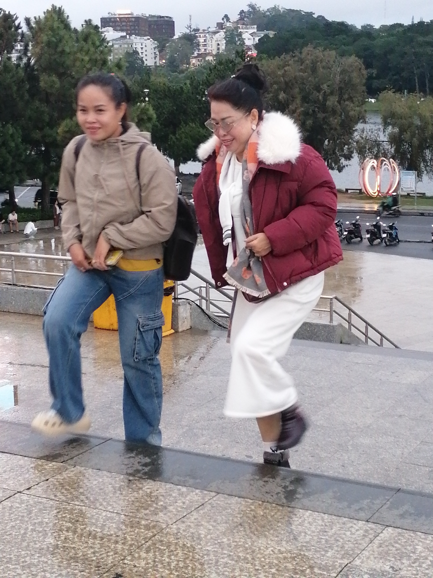 In Da Lat, Vietnam, on a wet, overcast day, two women are walking down an outdoor staircase. The woman on the left wears a tan hooded jacket, a yellow belt, and light blue cargo pants, with beige clog-like shoes. She carries a black backpack. The woman on the right is dressed in a maroon puffer jacket with a white faux-fur collar, a white scarf, and white pants, with dark shoes and white socks. She appears to be wearing glasses. The background shows a body of water, trees, buildings, and several motorcycles parked on a street. The overall impression is of a casual, outdoor urban environment.