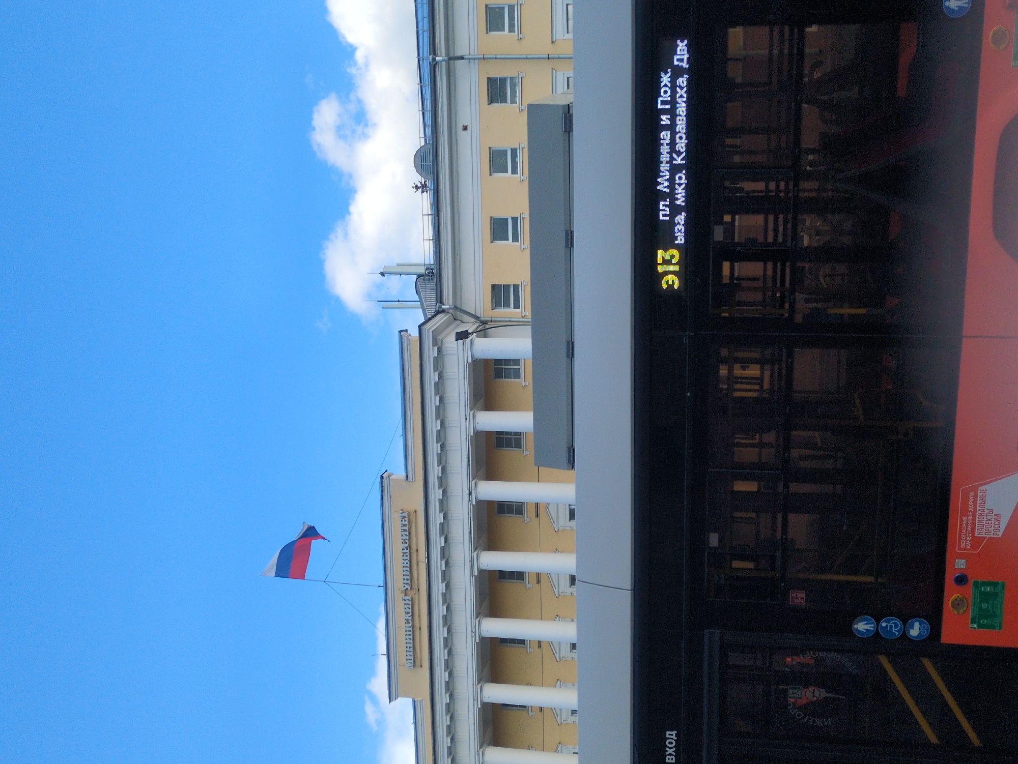 This media file captures a vertical, outdoor scene on a bright, clear day, indicated by the vibrant blue sky and scattered white clouds. The primary subjects are a building with classical architecture and a modern bus.
The building, likely a university as suggested by the text "МИНИНСКИЙ УНИВЕРСИТЕТ" (Mininsky University) visible on its facade, features rows of windows and prominent white columns supporting an upper story. A Russian flag, with its distinctive white, blue, and red horizontal stripes, is seen flying from a flagpole on the left side of the frame, adding a patriotic element to the setting.
In the foreground, and taking up a significant portion of the right side of the image, is a bus. Its dark windows reflect the surrounding environment, and a digital display board on the side of the bus shows route information in Russian, including the number "313" and destinations such as "пл. Минина и Пож." (Minina and Pozharsky Square) and "ул. Каравайха" (Karavaikha Street). Additional text on the bus, "БЕЗОПАСНЫЕ КАЧЕСТВЕННЫЕ ДОРОГИ НАЦИОНАЛЬНЫЕ ПРОЕКТЫ РОССИИ" (Safe High-Quality Roads National Projects of Russia), suggests it is part of a public transport initiative. The word "Вход" (Entrance) is also visible near the bus doors, indicating a point of access.
The overall impression is of a typical urban scene in Nizhny Novgorod, Russia, with a blend of historical architecture and modern transportation. The bright daylight suggests the photograph was taken during the daytime.