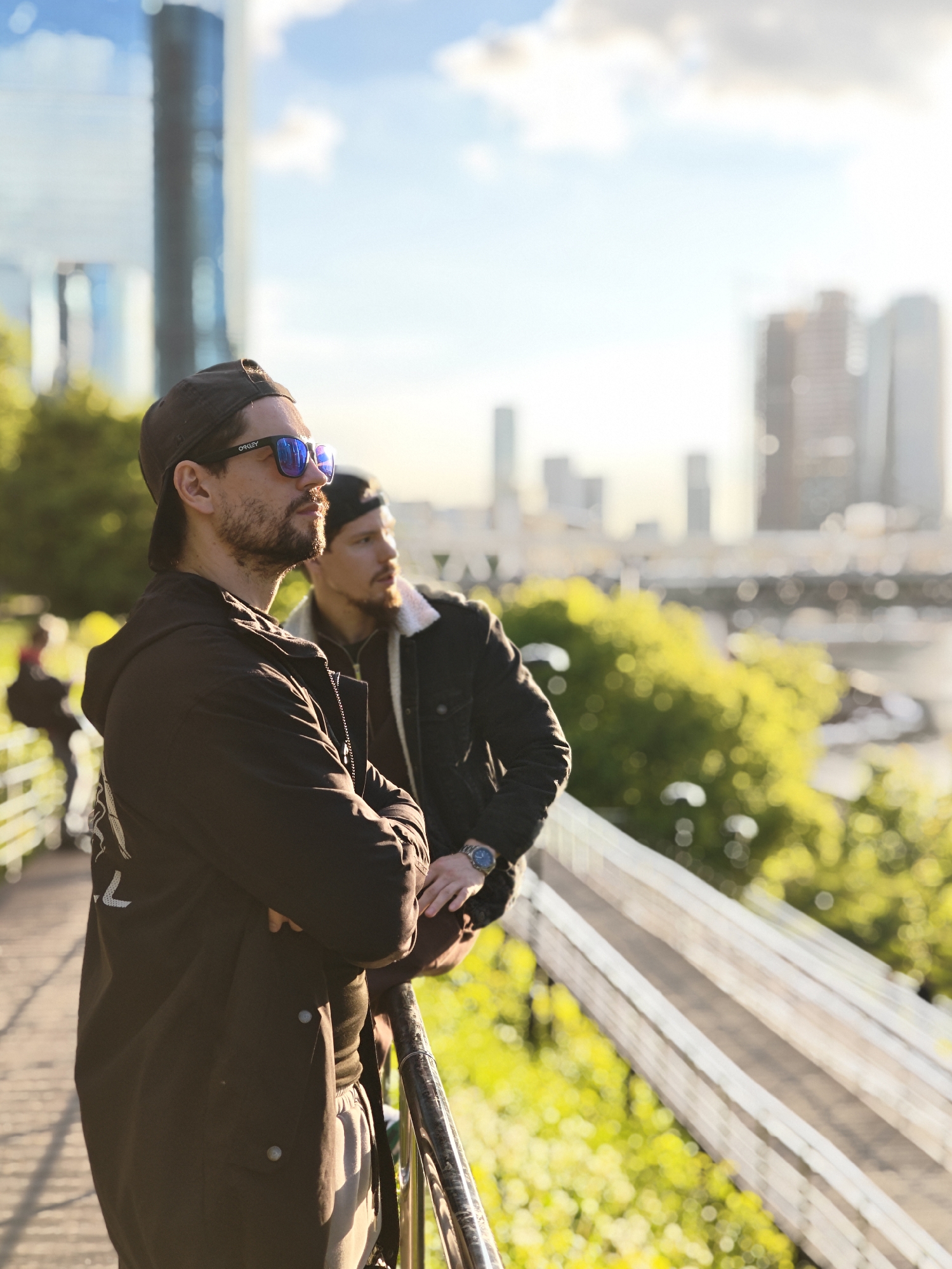 Two men stand on a walkway, leaning against a railing, looking out towards a blurred cityscape. The man in the foreground wears a dark cap, sunglasses, and a dark coat with arms crossed. The man behind him wears a jacket and has a beard. In the background is a blurred cityscape with tall buildings, and green foliage. The location is the Patriarshy Bridge (Патриарший мост) overlooking Moskva River in Moscow, Russia.