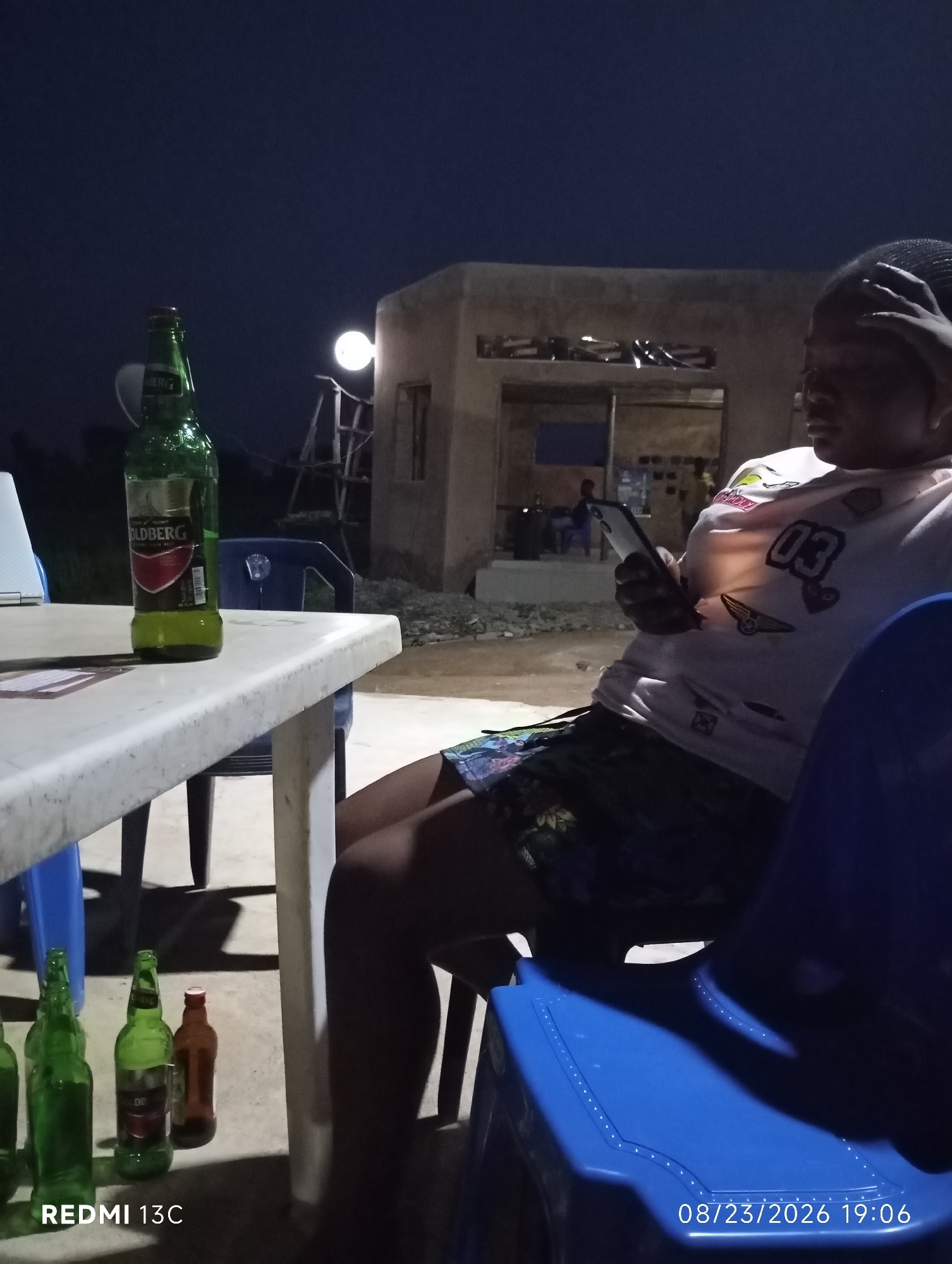 The image depicts a nighttime scene at an outdoor establishment in Shendam, Nigeria. A person is seated in a blue chair, holding a smartphone and wearing a light-colored t-shirt with graphic designs, including the number "03". In the foreground, on a white table, are several green glass beer bottles, one of which is a "Goldberg" brand. Another brown bottle is also present. The background reveals a simple, unfinished building with a light illuminating an opening. The overall atmosphere suggests a casual gathering or relaxation spot.