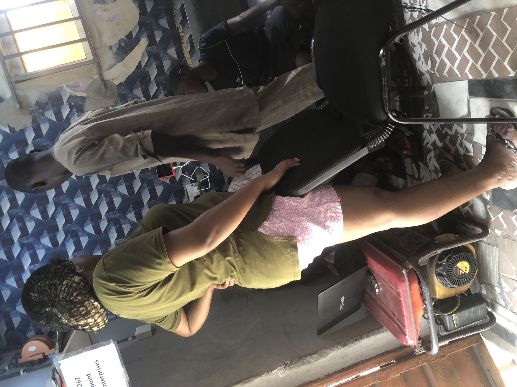 In Ado, Nigeria, an indoor scene shows two individuals, a woman in a green t-shirt and pink patterned shorts, and a man in a beige kaftan. The woman, partially obscured, stands with her hand on her hip, looking away from the camera. She wears a hair net covering her braided hair. The man, seen from the back, faces her and appears to be talking. To the right, a third person, a young boy in a dark t-shirt and shorts, sits on a couch, looking to the left. In the foreground, a red and yellow generator is visible, with a black laptop placed on top of it. The room has patterned blue wallpaper and a window with metal bars. The floor is tiled with a diamond pattern in brown and white. The overall impression is of a casual, everyday setting in a Nigerian urban environment.