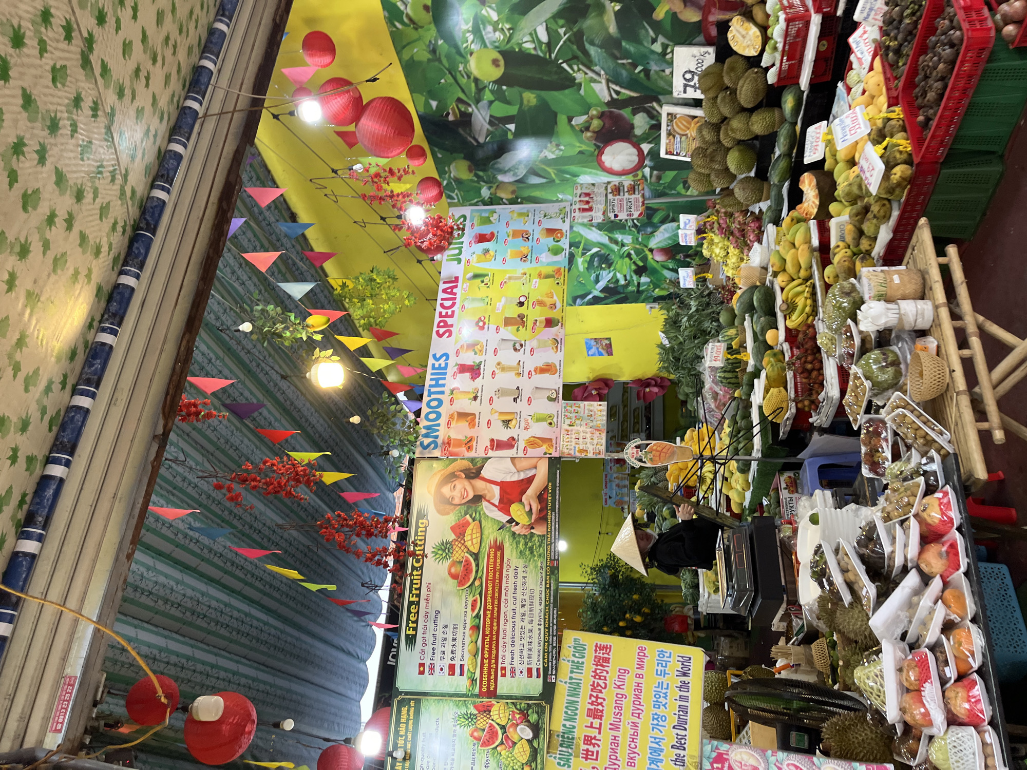 This is a vibrant, eye-level, medium-wide shot of a fruit stall in Nha Trang, Vietnam. The stall is located on a street, likely a market, under a striped awning decorated with red lanterns and colorful triangular flags. The background is a yellow wall adorned with a large mural of lush green leaves and fruit.
In the foreground, a variety of fruits are neatly displayed in plastic containers and baskets on tables. These include durian, mangosteen, lychees, and other tropical fruits. A woman wearing a traditional conical hat stands in the middle of the scene, appearing to be attending the stall. She is holding a tool and is positioned behind a display of fruit. To the left, a large yellow sign is visible, advertising "the Best Durian in the World" in multiple languages. To the right, a menu board with images of smoothies and special juices is prominently displayed.
The lighting is bright, illuminating the colorful fruits and decorations. The overall impression is one of a bustling and well-stocked market stall offering fresh produce and beverages.