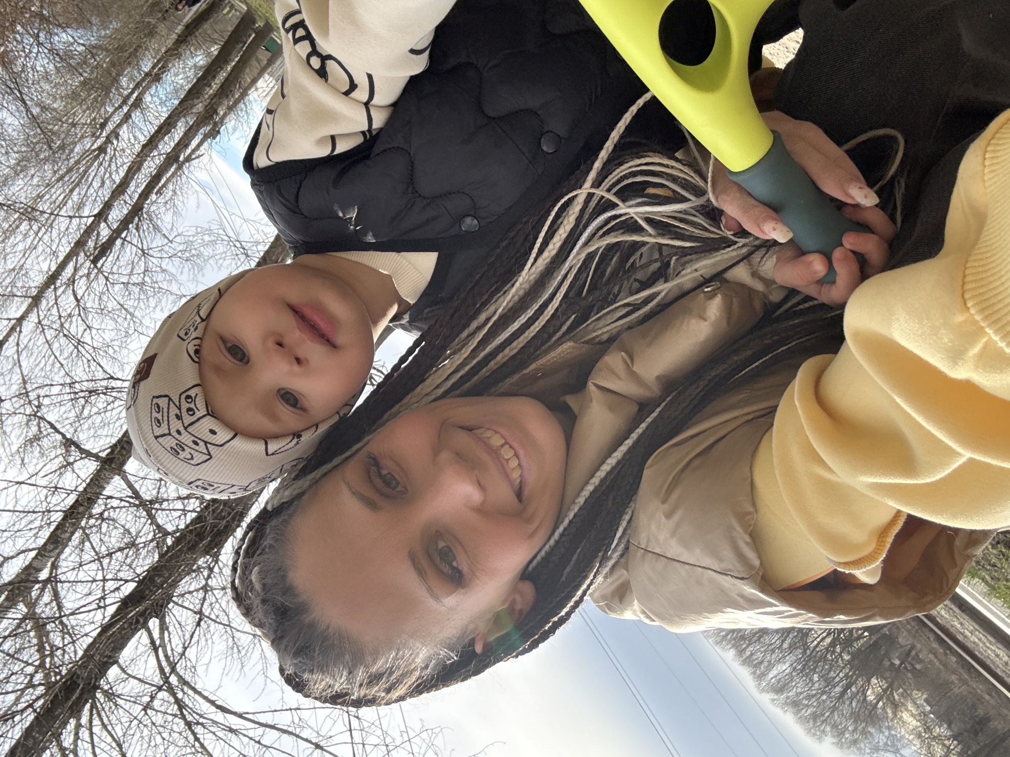 A woman and a baby are positioned in front of a background of bare trees and sky, with a power line visible. The woman has brown hair with blonde extensions, wears a tan jacket over a yellow sweater, and smiles at the camera. She holds a green and gray object, possibly a toy or tool. The baby wears a patterned hat, a black vest over a light-colored sweater, and looks toward the camera with a neutral expression. The location is outdoors, near trees, possibly in Fedorino, Russia.