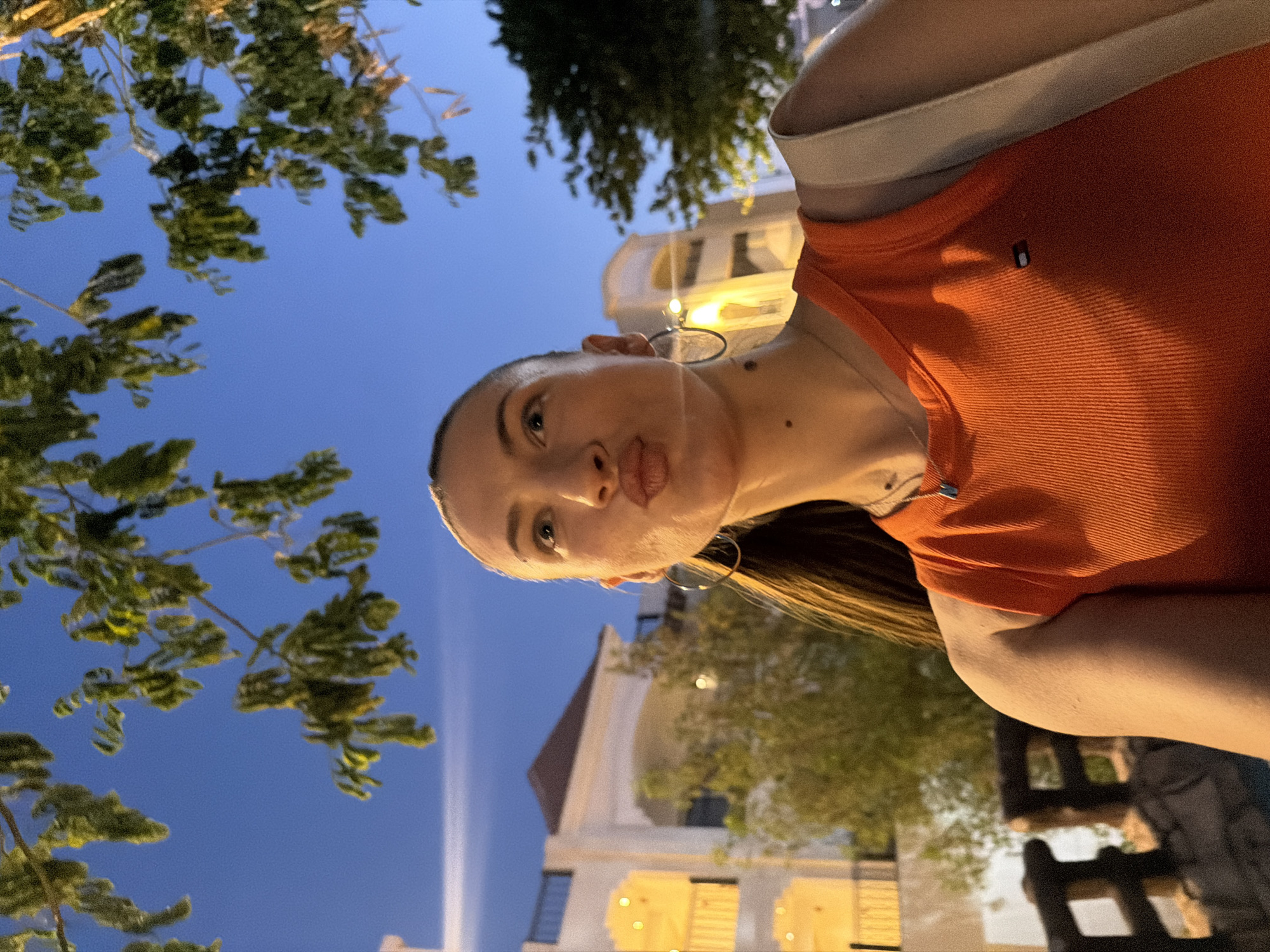 A young Caucasian woman with blonde hair pulled back and a pouty expression is taking a selfie outdoors at dusk, in Sharm El Sheikh, Egypt. She is wearing an orange ribbed tank top with a small Tommy Hilfiger logo on the chest and a delicate silver necklace. Large hoop earrings adorn her ears. She is standing in front of a building with arched windows and warm yellow lighting, possibly a hotel or restaurant. Trees with green foliage are visible overhead, silhouetted against a clear blue sky. A brown wooden railing is in the foreground to her left. The overall atmosphere suggests a casual evening outing in a resort area.