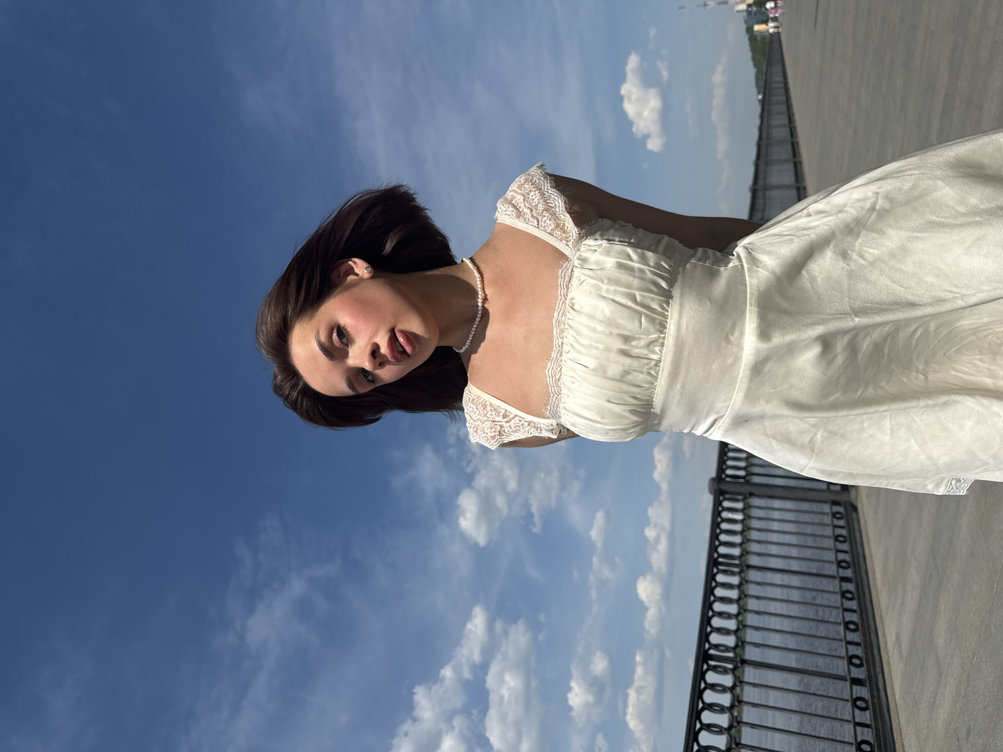 A young woman with dark hair and fair skin is posing on a city bridge in Perm, Russia. She is wearing a white dress with lace trim and a pearl necklace. She is leaning back, looking directly at the camera with a slight smile. The background is a blue sky with some clouds and a metal railing along the side of the bridge. The bridge is made of concrete. There are some buildings visible in the far distance. The image appears to have been taken during the daytime.