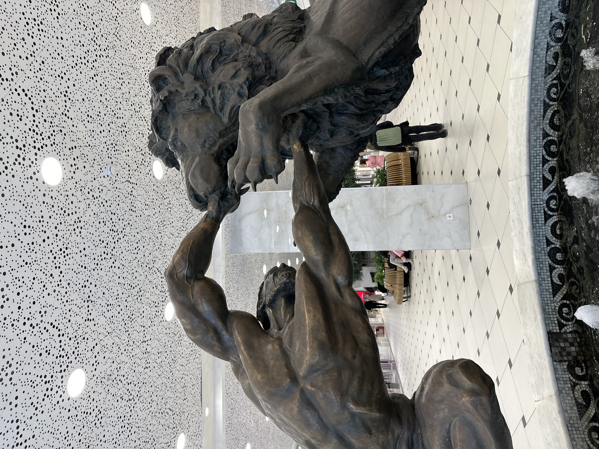 The image captures a bronze statue depicting a muscular male figure wrestling or lifting a lion. The man, seen from the back and side, has well-defined muscles, particularly in his arms and shoulders, as he holds up the powerful lion. The lion, also rendered in bronze, is detailed with a flowing mane and prominent claws on its paws, which are visible near the man's hands.
The scene is set in a modern indoor public space, likely a shopping mall or large hall. The ceiling is a striking feature, uniformly white with numerous small, irregularly distributed black circular perforations, and illuminated by several bright, round recessed lights. The floor is a light-colored tiled surface with a diagonal grid pattern, accentuated by small black square tiles at the intersections, forming a diamond design.
To the right, a decorative fountain is visible, featuring dark water and an intricate mosaic border with black and white swirling patterns. Two small water jets spray upwards from the fountain. In the background, several individuals can be seen on the tiled floor, some standing and others seated on long, light-colored benches. One person stands in dark clothing with a light green shirt. White marble panels are integrated into the architecture, appearing on low walls and structural elements. The overall impression is a well-lit, contemporary interior. No visible text is present.