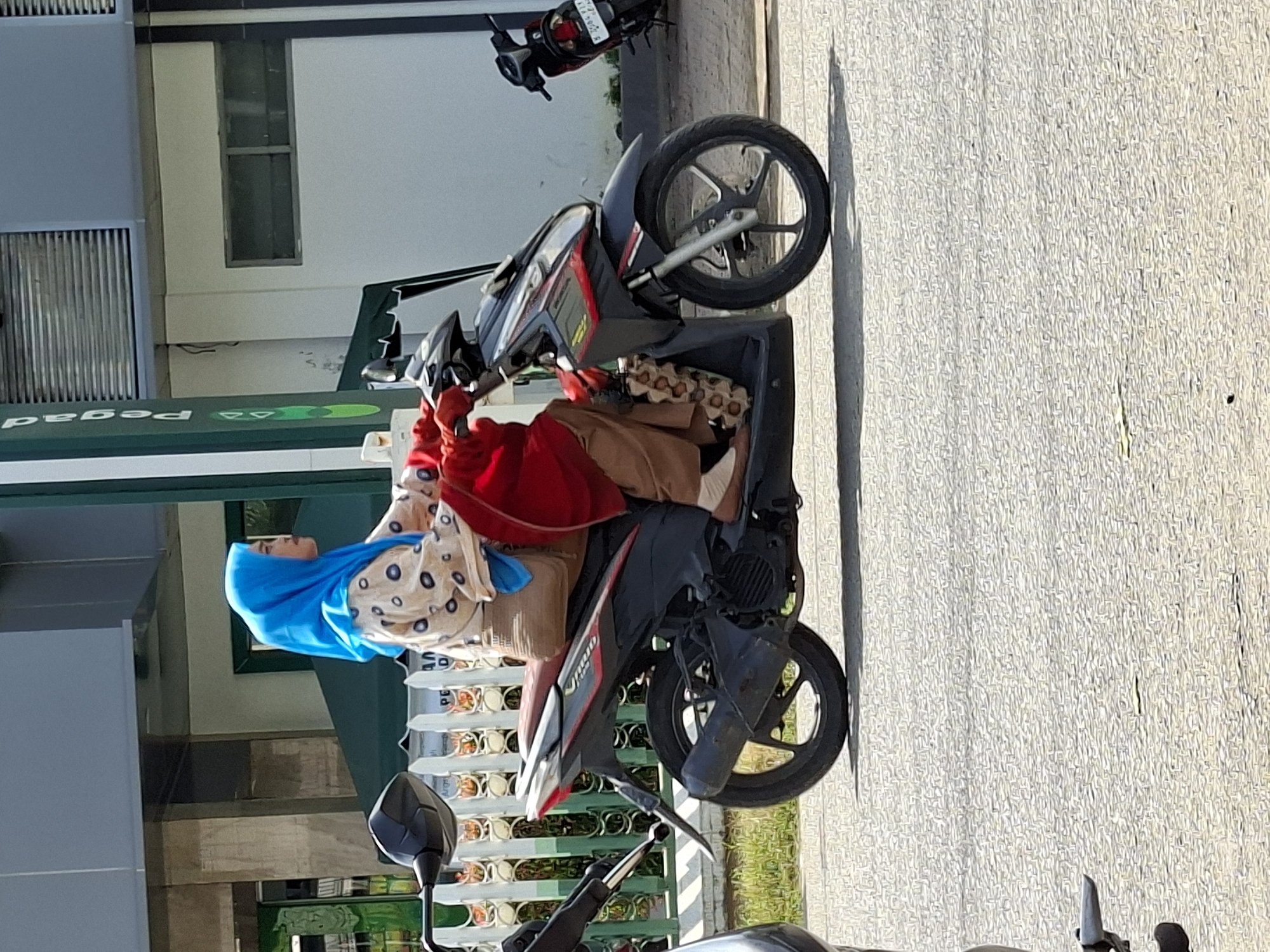 A woman wearing a bright blue hijab and a patterned shirt rides a black and red scooter. She is also wearing brown pants and carrying a red shawl draped over her arm. The scooter has a visible exhaust pipe and a brown seat. Behind the woman, a white picket fence borders a grassy area. In the background, a building with gray and white sections and a green awning is visible, along with a vertical green sign with white lettering that reads "Pegadaian". The scene appears to be on a paved road in Lhokseumawe, Indonesia.