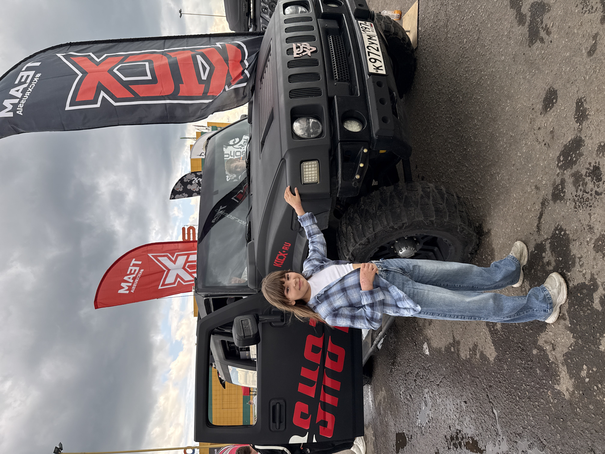 The image shows a black Hummer H2 parked in Rusanovo, Russia. The license plate is visible (K 972 YM 197). The car is branded with "KICK.RU" and "TEAM KICKX" graphics. A young person, wearing a blue and white plaid shirt and jeans, is leaning against the Hummer. The driver-side door of the vehicle is open. The sky is overcast. The ground appears to be a paved surface with some wet areas.