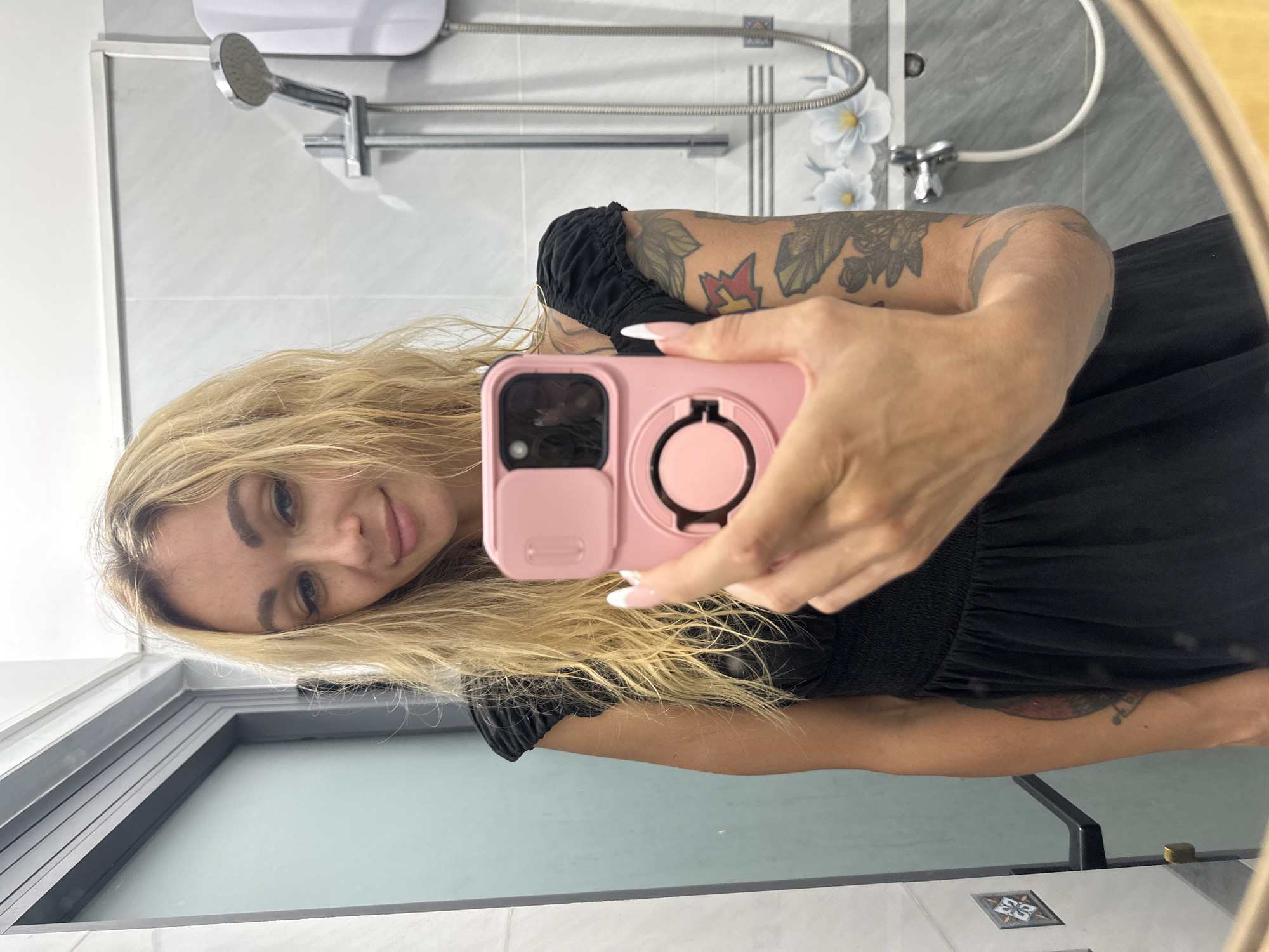 This image captures a mirror selfie of a blonde woman inside a modern bathroom, located in Mũi Né, Vietnam. The woman, with long, wavy blonde hair, gazes directly into the camera with a subtle, gentle smile. She appears to be wearing a black top or dress with distinctive ruched sleeves. Her arms are notably adorned with multiple tattoos, featuring various designs including floral patterns and a vibrant red and yellow flame-like symbol on her upper arm. Her fingernails exhibit a classic French manicure.
She is holding a pink smartphone, equipped with a prominent circular ring holder or stand on its back, which she uses to take the photo.
The background, reflected in the mirror, reveals a contemporary bathroom setting. The walls are tiled in a combination of light grey and darker grey, with the darker tiles featuring an elegant white floral pattern. Visible fixtures include a wall-mounted silver shower head, a flexible shower hose, and a white electric water heater. A clear shower partition with a dark frame is also reflected. Near the bottom of the frame, a small decorative tile with a geometric design is visible, likely part of the floor or a lower wall. There is no explicit text visible within the image. The scene conveys a casual, private moment.