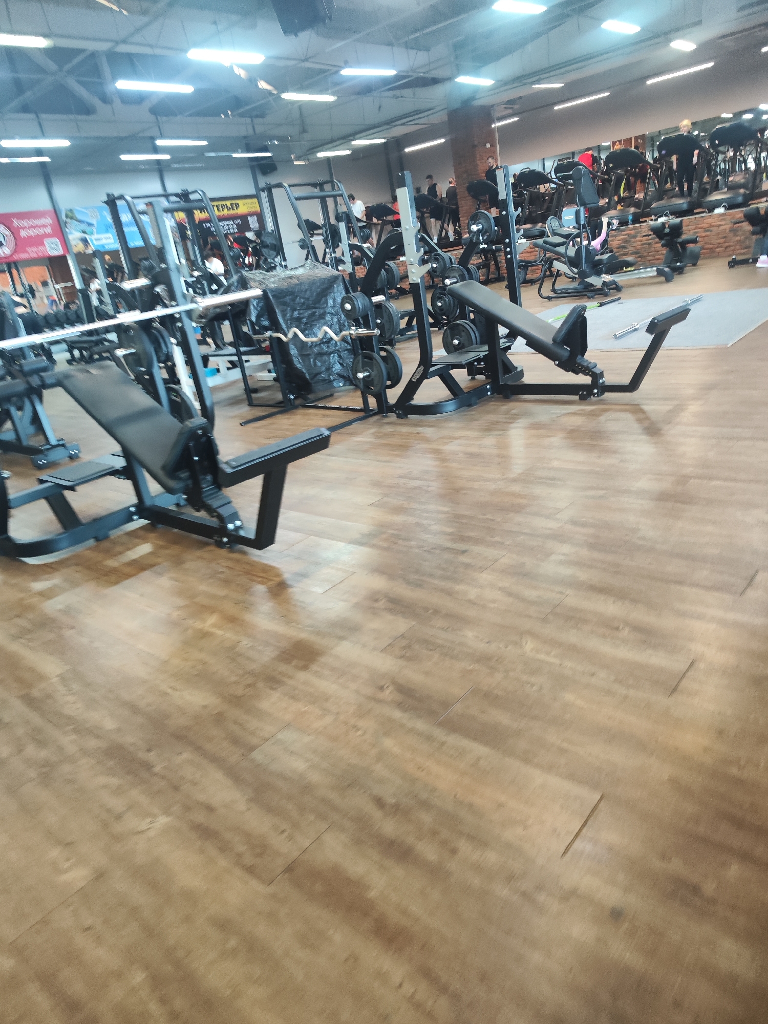 A gym in Chaikovsky, Russia, featuring various weightlifting machines, including seated chest press machines with attached weights. People are exercising in the background. The floor is made of light-colored planks. A machine is covered with a black tarp.