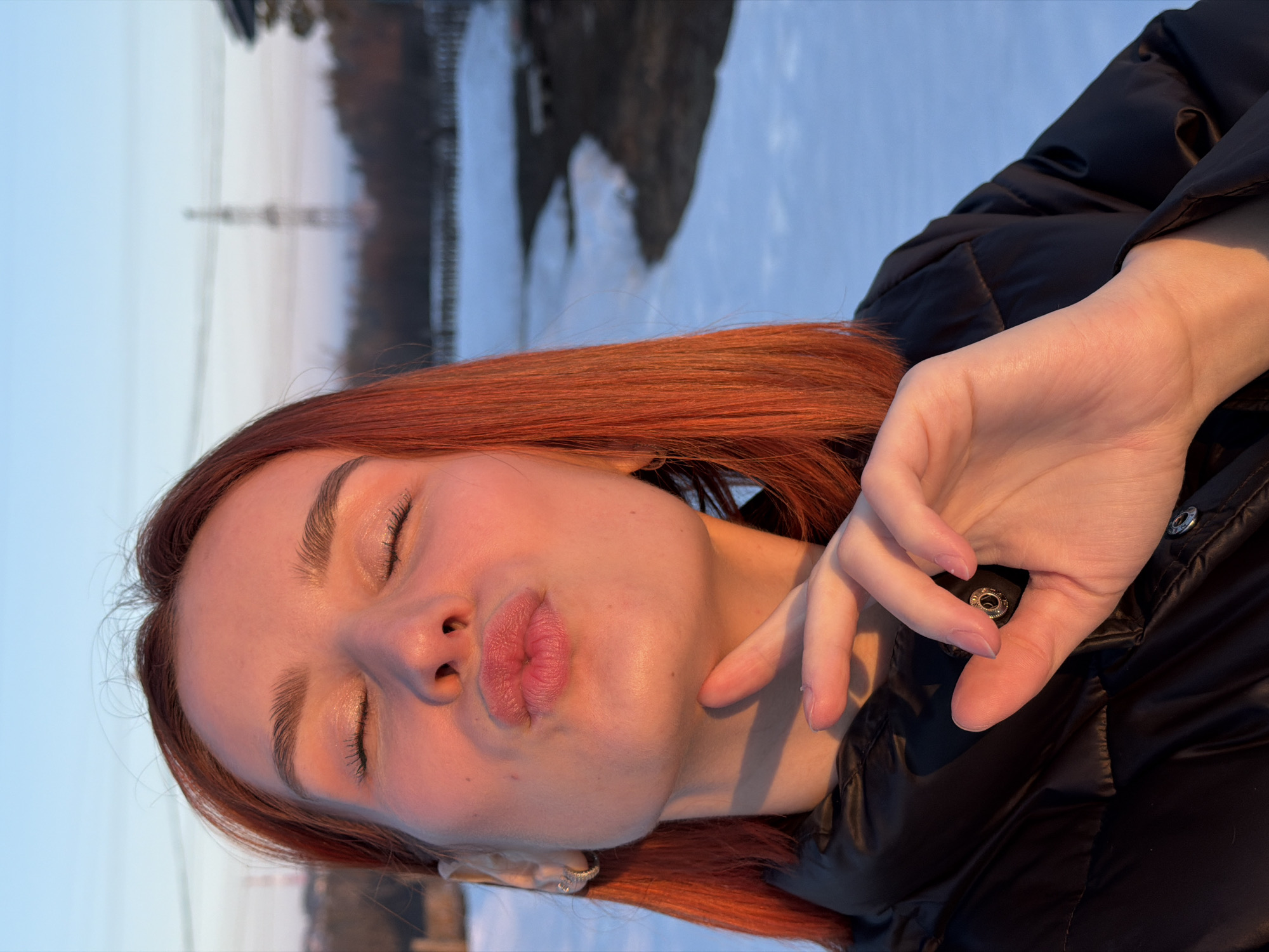 A close-up portrait in Chaikovsky, Russia, features a young female with reddish-orange hair and fair skin. Her eyes are closed, and her lips are pursed in a kissing gesture. She is wearing a black puffer jacket with visible snap buttons and a silver hoop earring in her left ear. Her right index finger is positioned against her chin. The scene is illuminated by warm, golden light, casting soft shadows. The background, out of focus, displays a snow-covered ground with patches of exposed dark earth, distant bare trees, and a prominent metal power transmission tower against a clear sky transitioning from light blue to a pale, warm orange near the horizon.